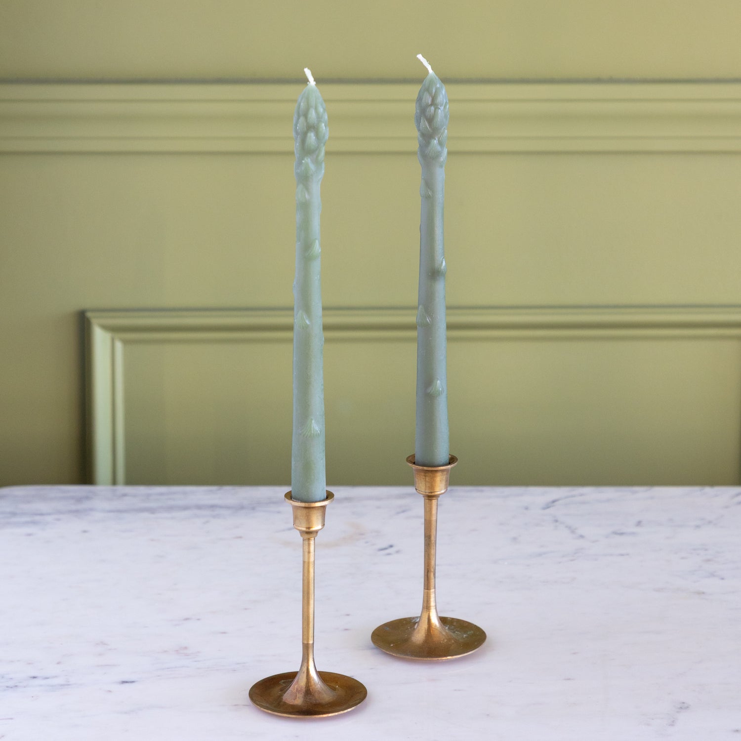 Two Asparagus Shaped Taper Candles in gold candle holders on a marble table.