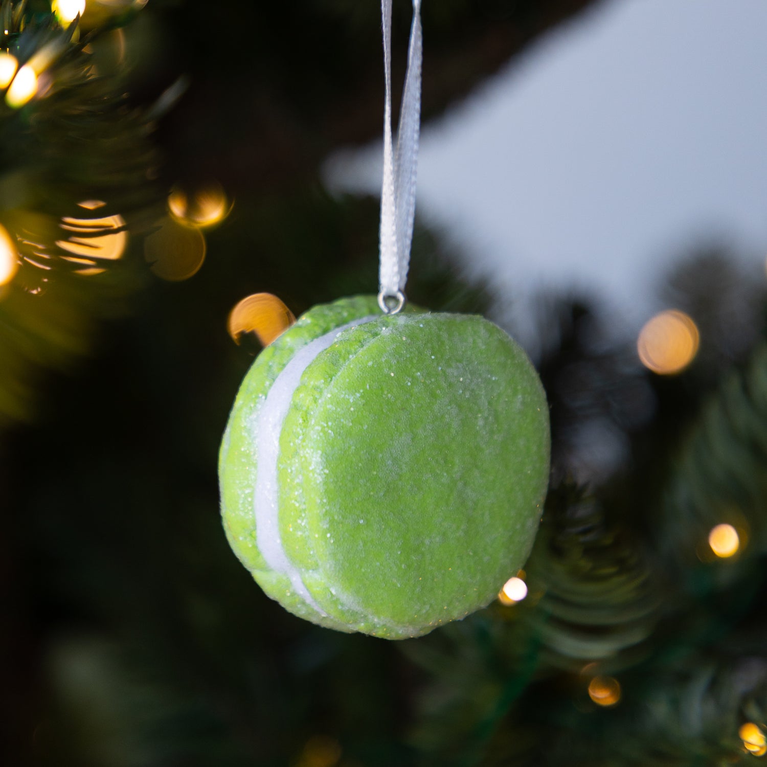 Green macaron ornament hanging on a Christmas tree with blurred lights in the background.
