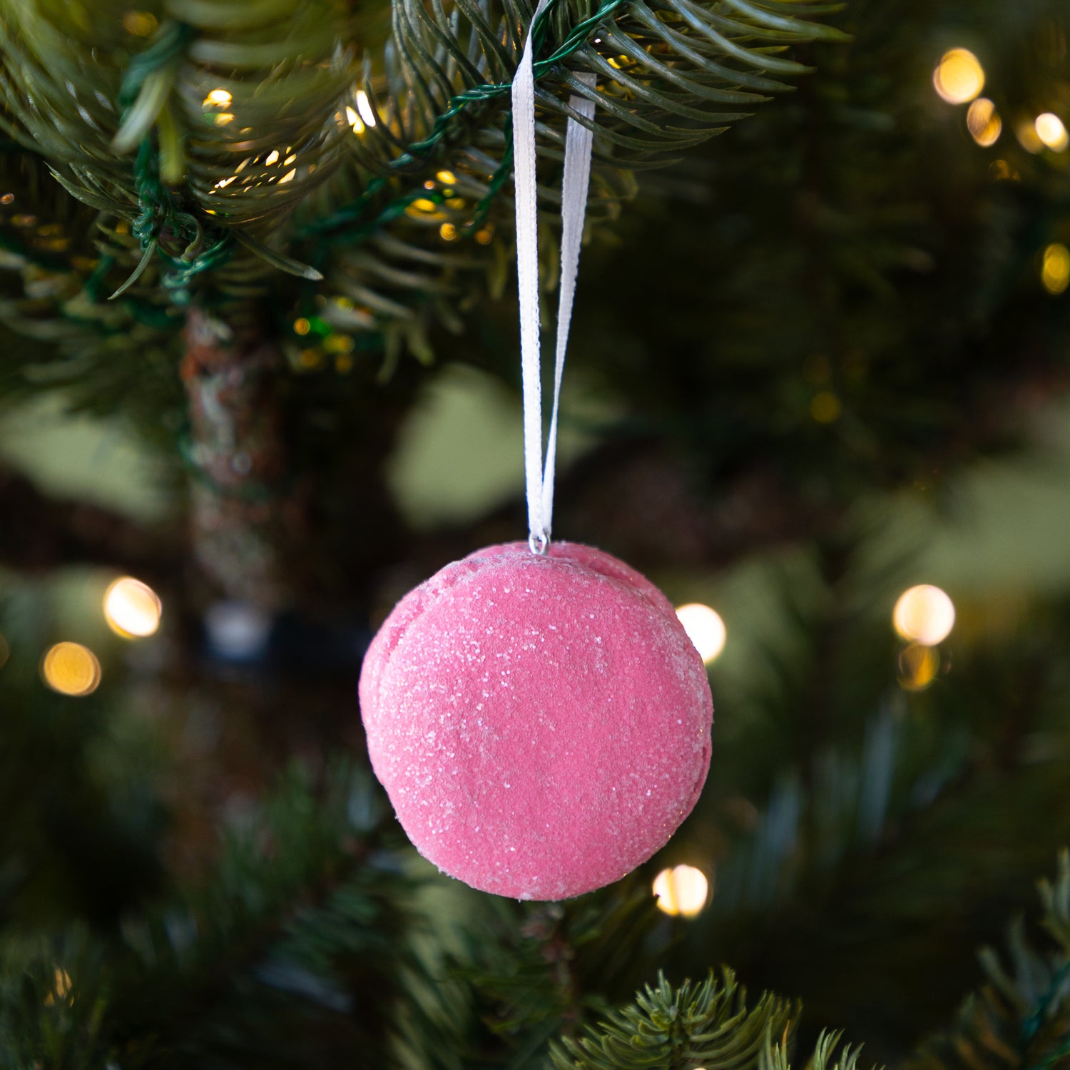 Pink macaron ornament hanging on a Christmas tree with blurred lights in the background.