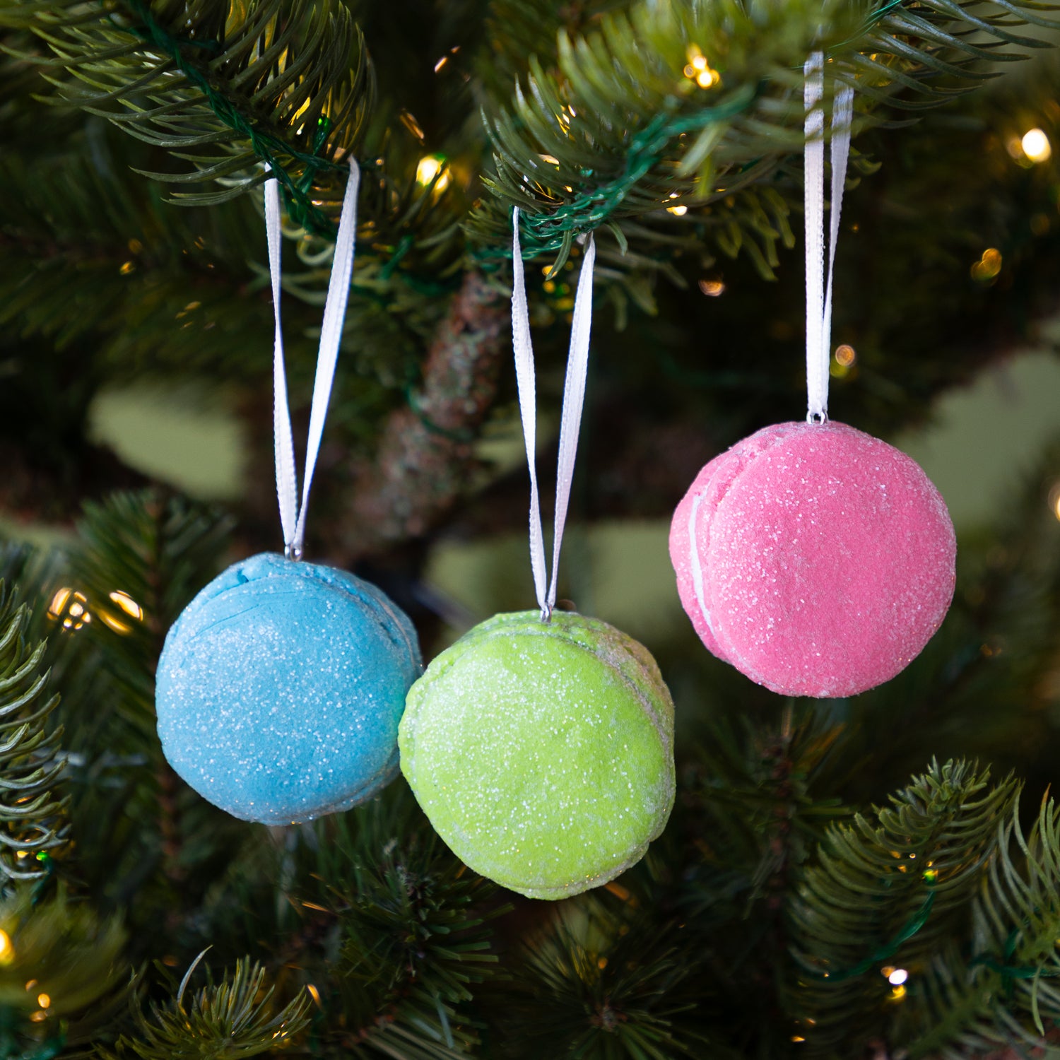 Three macaron-shaped ornaments in blue, green, and pink hanging on a Christmas tree.