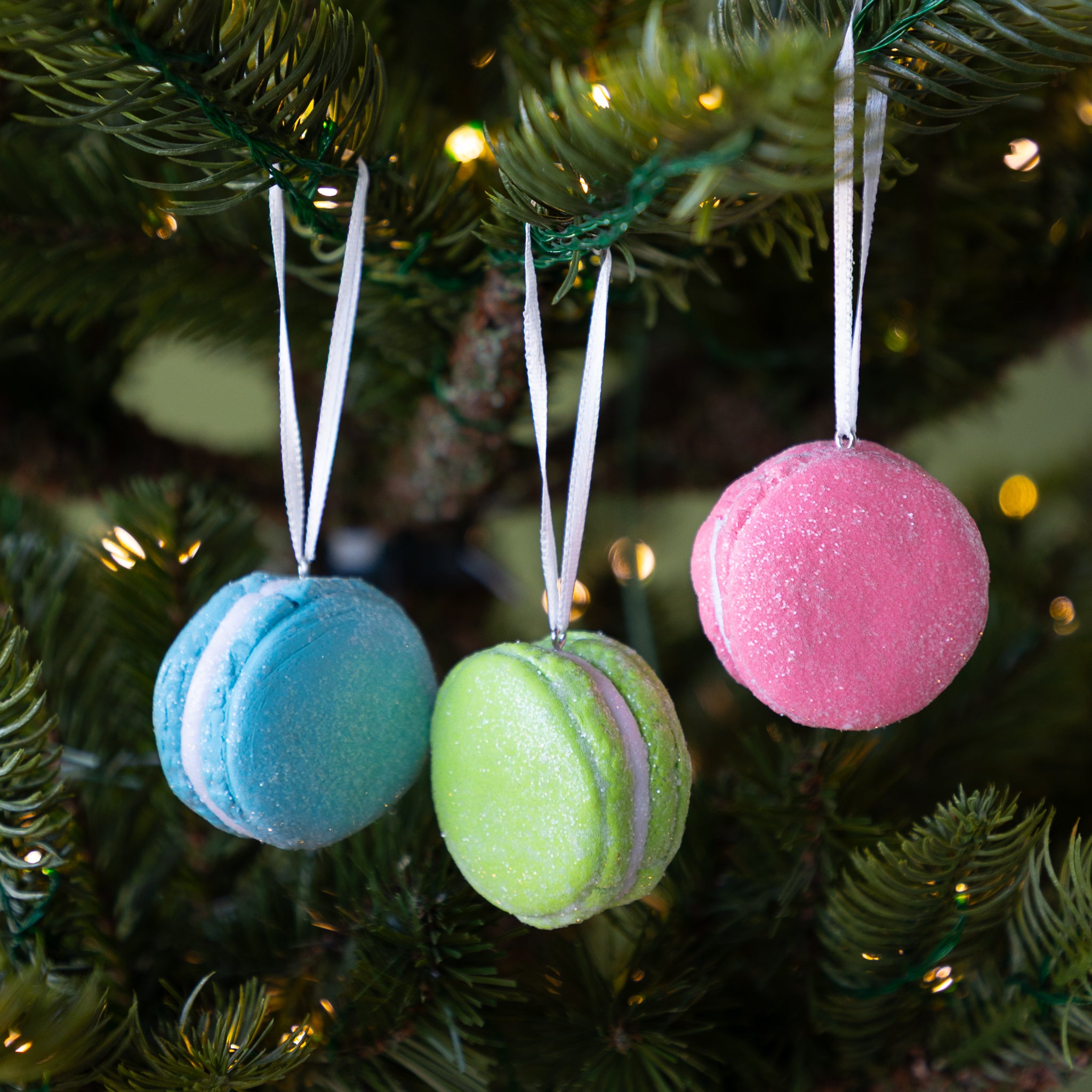 Three macaron-shaped ornaments in blue, green, and pink hanging on a Christmas tree.