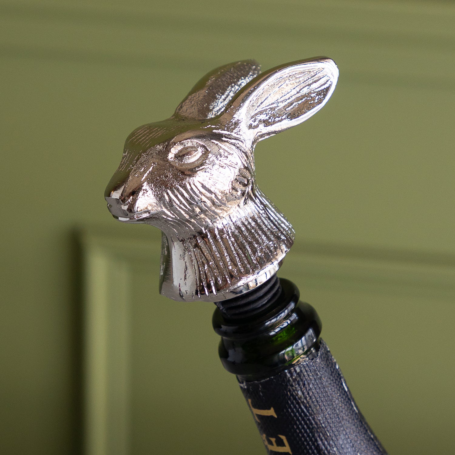 A close-up of a metallic silver rabbit-shaped bottle stopper inserted into the neck of a dark glass wine bottle, set against a soft olive green paneled background.
