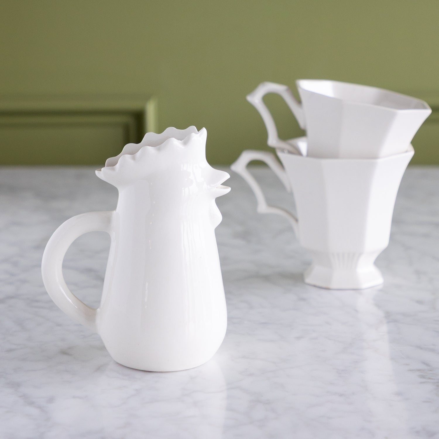 Stoneware Chicken Creamer and stacked cups on a marble table with a green wall backdrop.