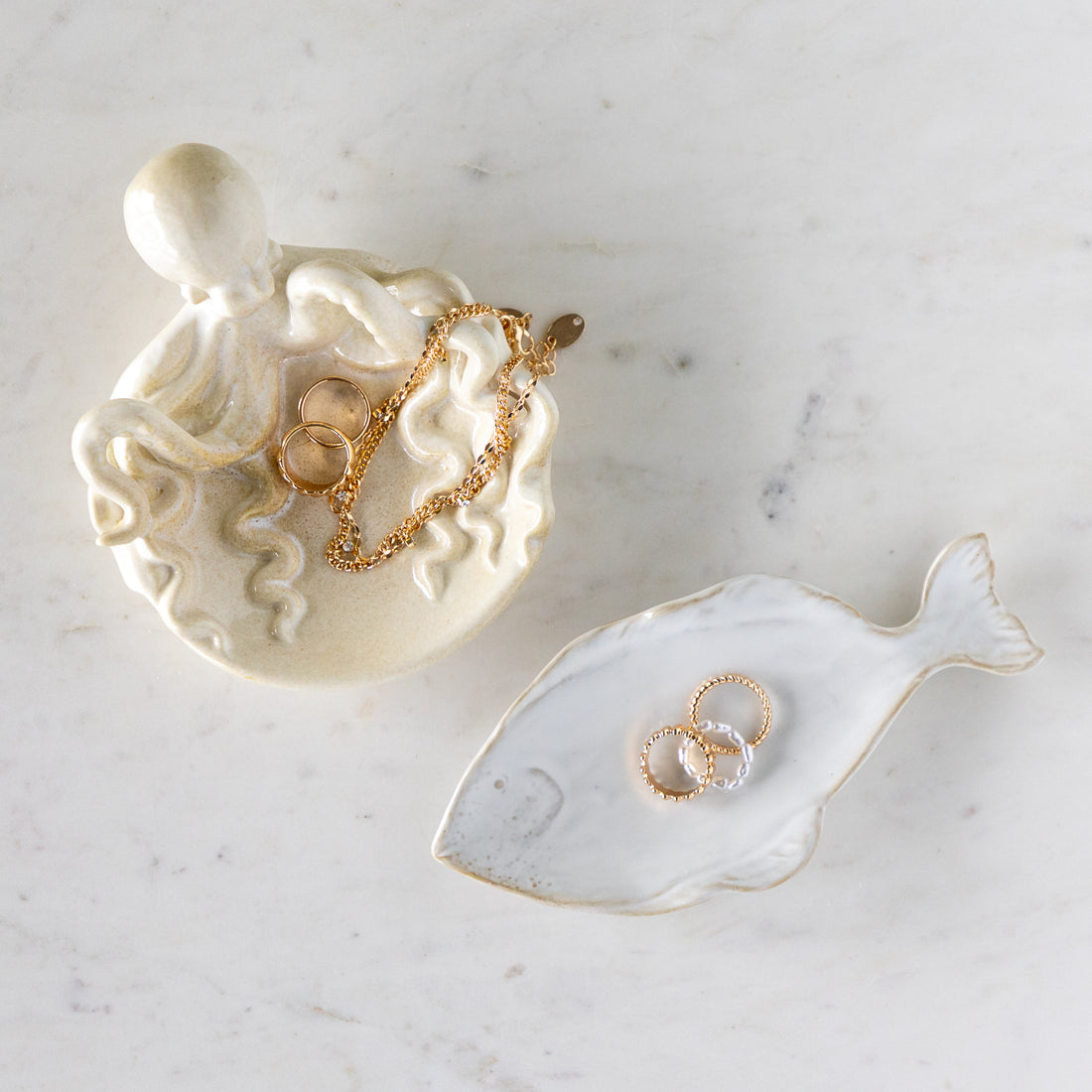 Stoneware Fish Shaped Plate with rings in it, next to Stoneware Octopus Dish filled with rings and a necklace, on a marble table.