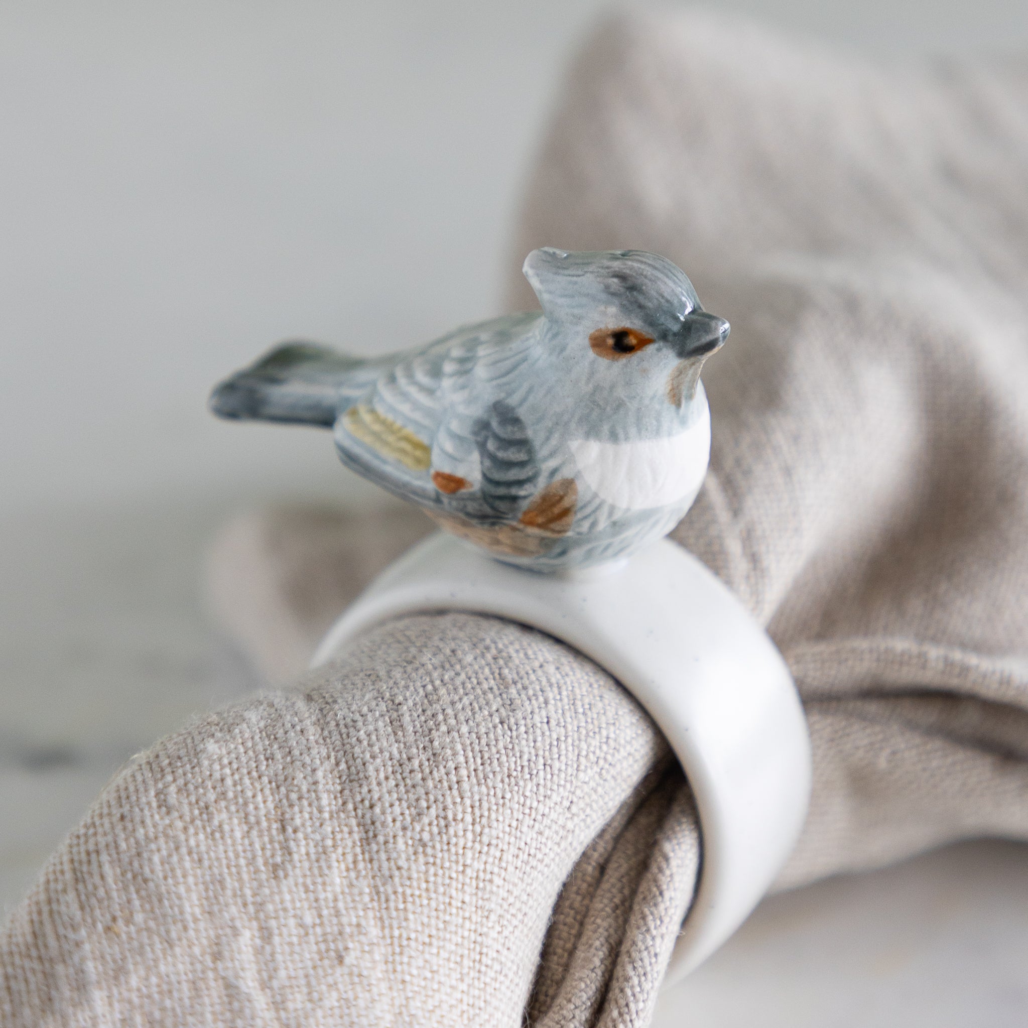 Napkin ring with grayish-blue bird design around a folded beige napkin against a neutral background.