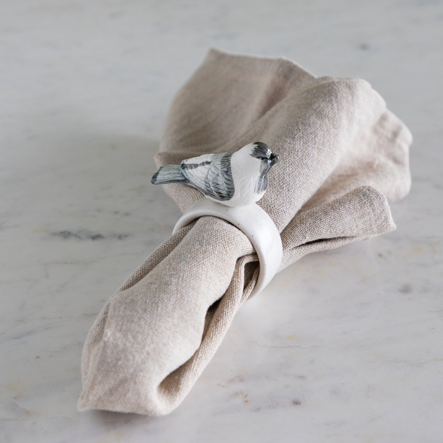 Napkin ring with white and gray bird design around a folded beige napkin against a neutral background.
