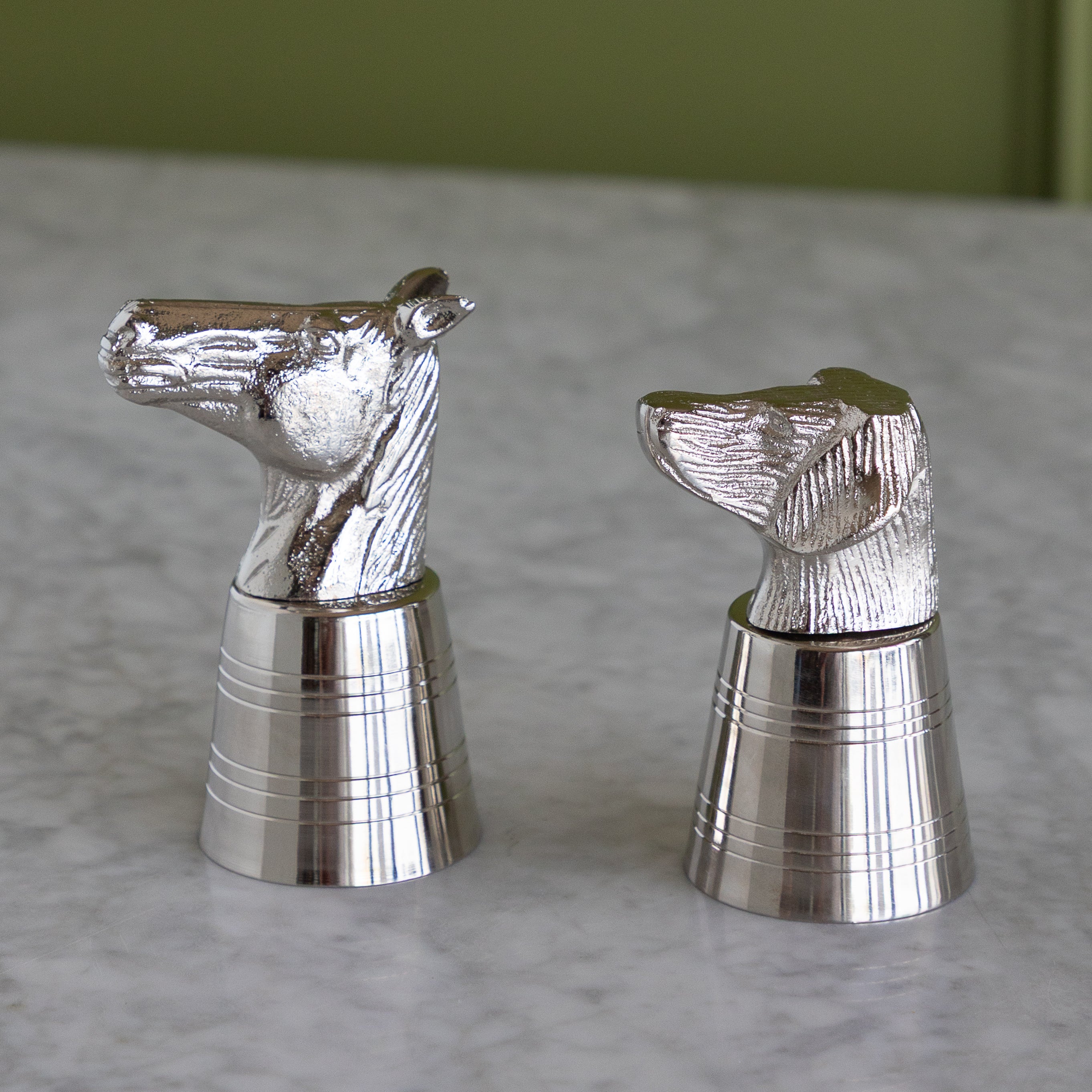 Metallic horse-shaped and dog-shaped jiggers with silver bases, displayed on a light marble surface with a soft-focus olive green background.