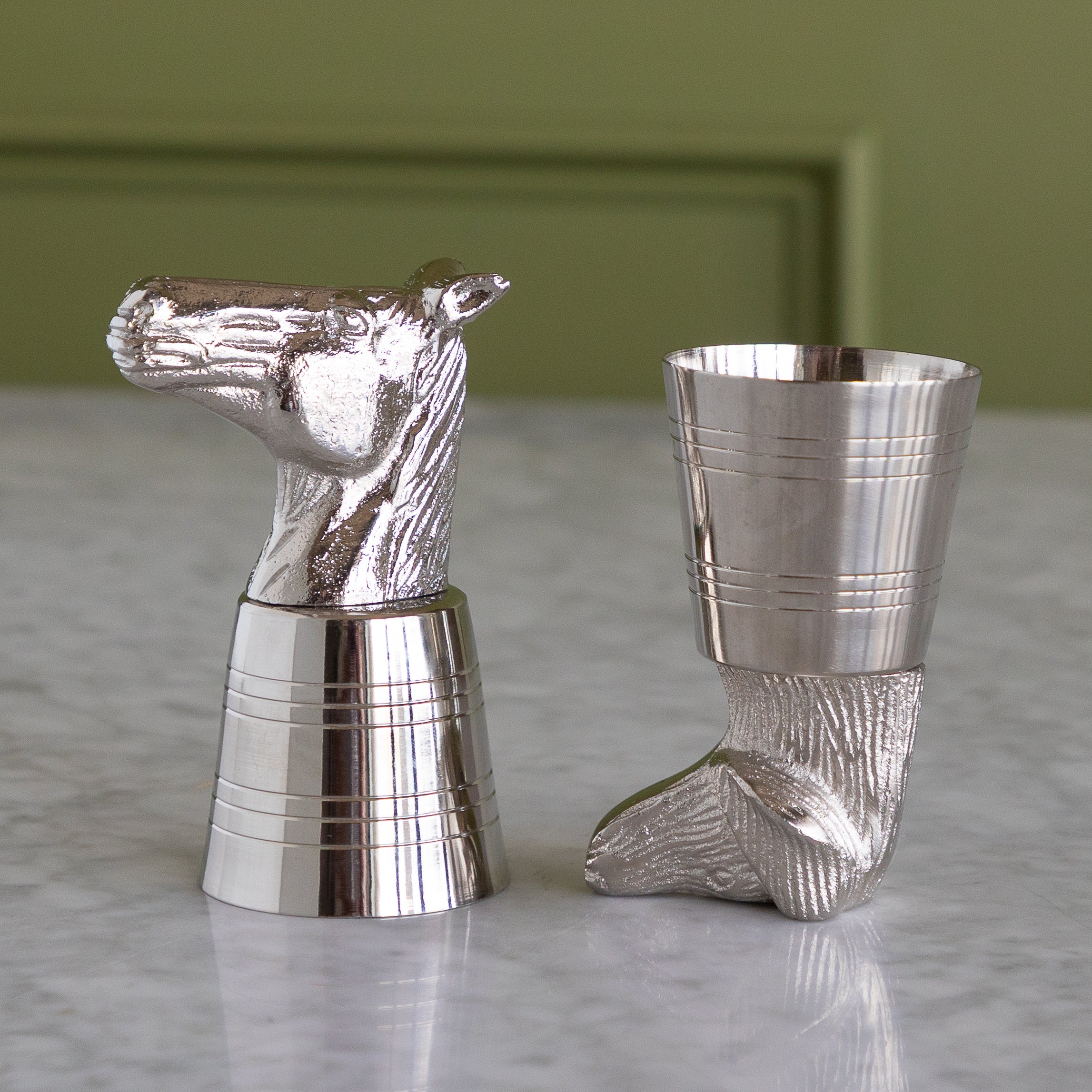 Two polished silver-toned metal shot glasses shaped like a horse head and a dog head displayed on a light marble surface with a soft olive green background.