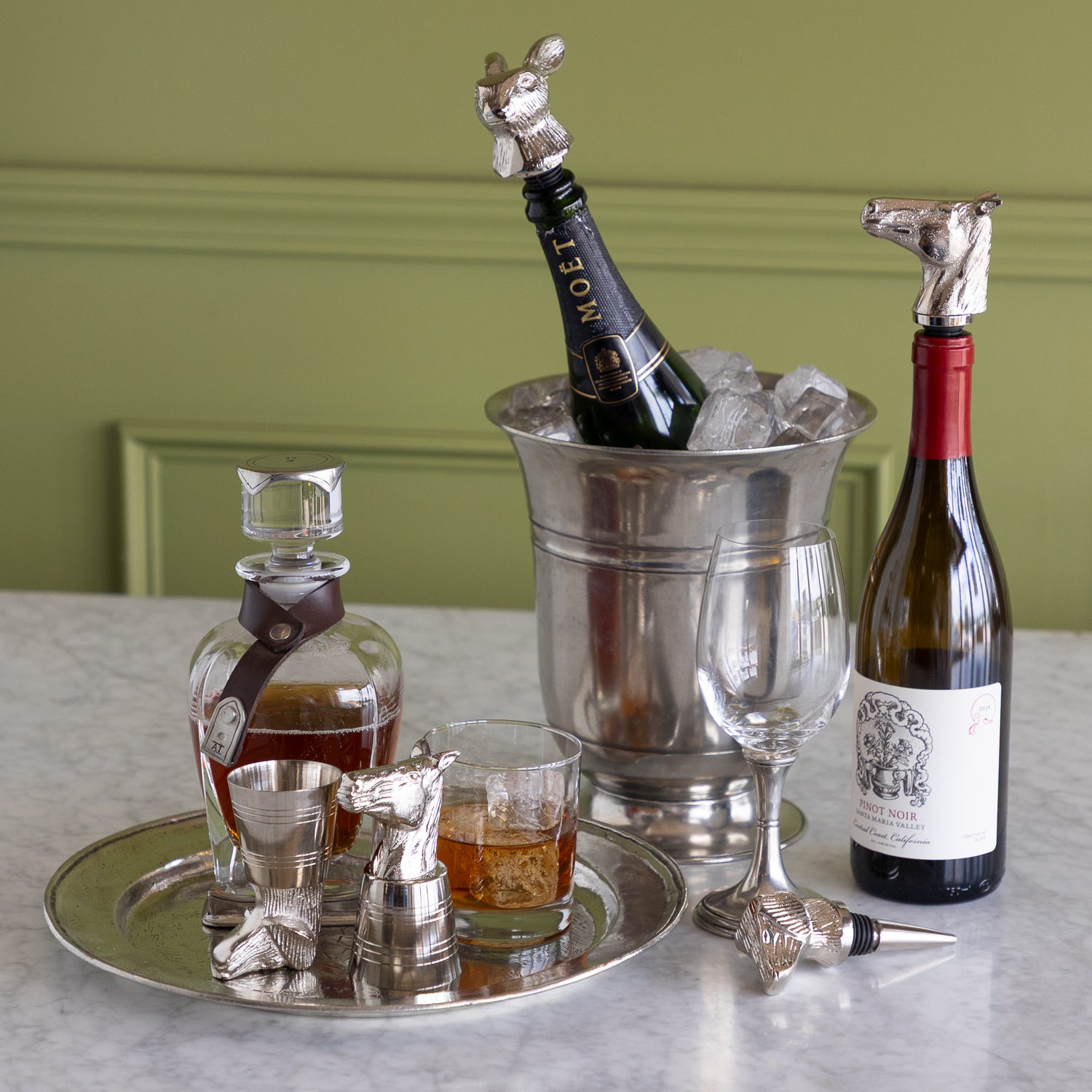 A styled bar scene on a marble countertop with a silver ice bucket holding a dark bottle, a wine bottle, a decanter, a filled coupe glass, a filled rocks glass, and several silver animal-shaped pourers and stoppers. A green paneled wall is in the background.