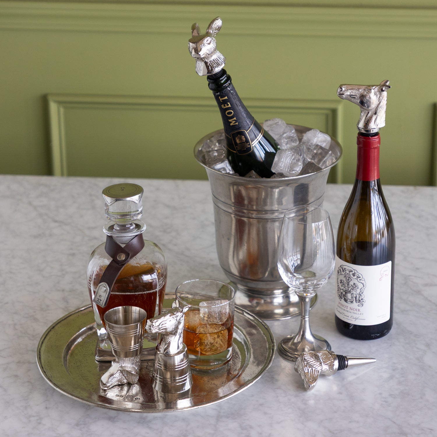 A styled bar setup on a light marble countertop against a green paneled wall, featuring a silver horse-shaped bottle stopper with a Moët &amp; Chandon champagne bottle in an ice bucket, a wine bottle with a horse stopper, a decanter, a jigger, a glass, and a wine glass.