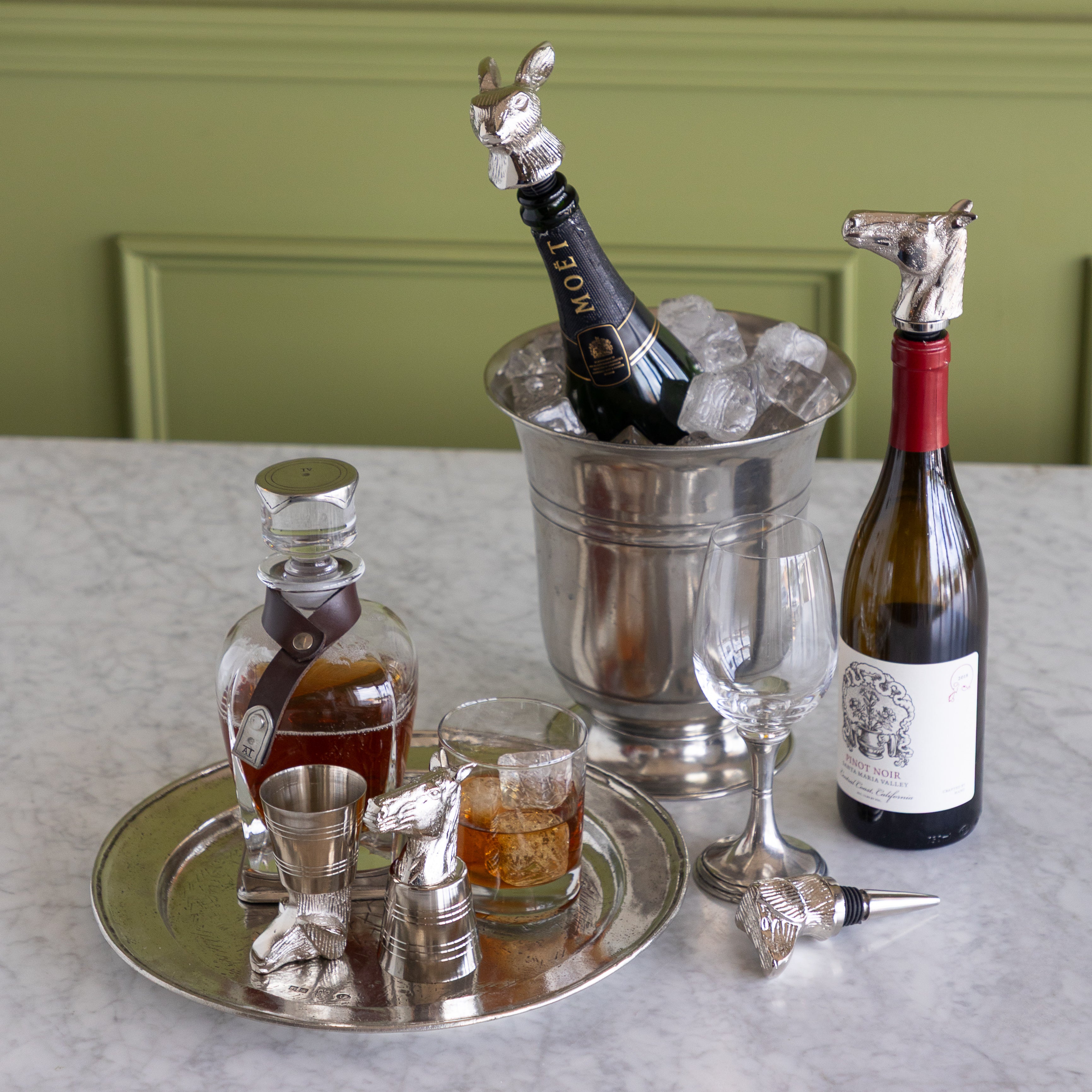 A styled bar setup on a light marble countertop against a green paneled wall, featuring a silver horse-shaped bottle stopper with a Moët &amp; Chandon champagne bottle in an ice bucket, a wine bottle with a horse stopper, a decanter, a jigger, a glass, and a wine glass.