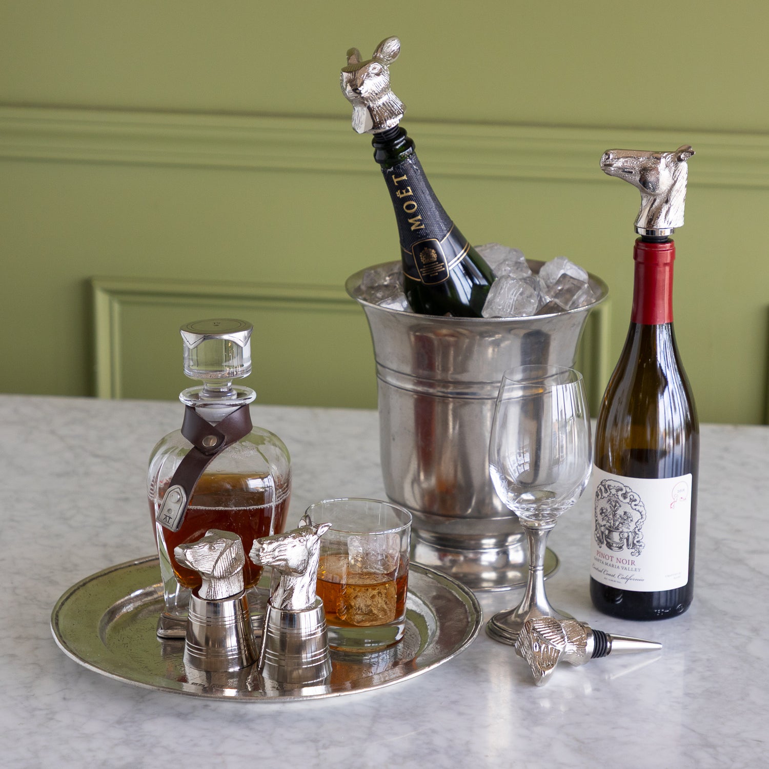 A styled bar scene on a marble countertop with a silver tray holding a decanter, a small glass, and two silver horse-shaped stoppers. A metal ice bucket with a Moet &amp; Chandon bottle and ice sits beside a wine bottle with a silver horse-shaped stopper.