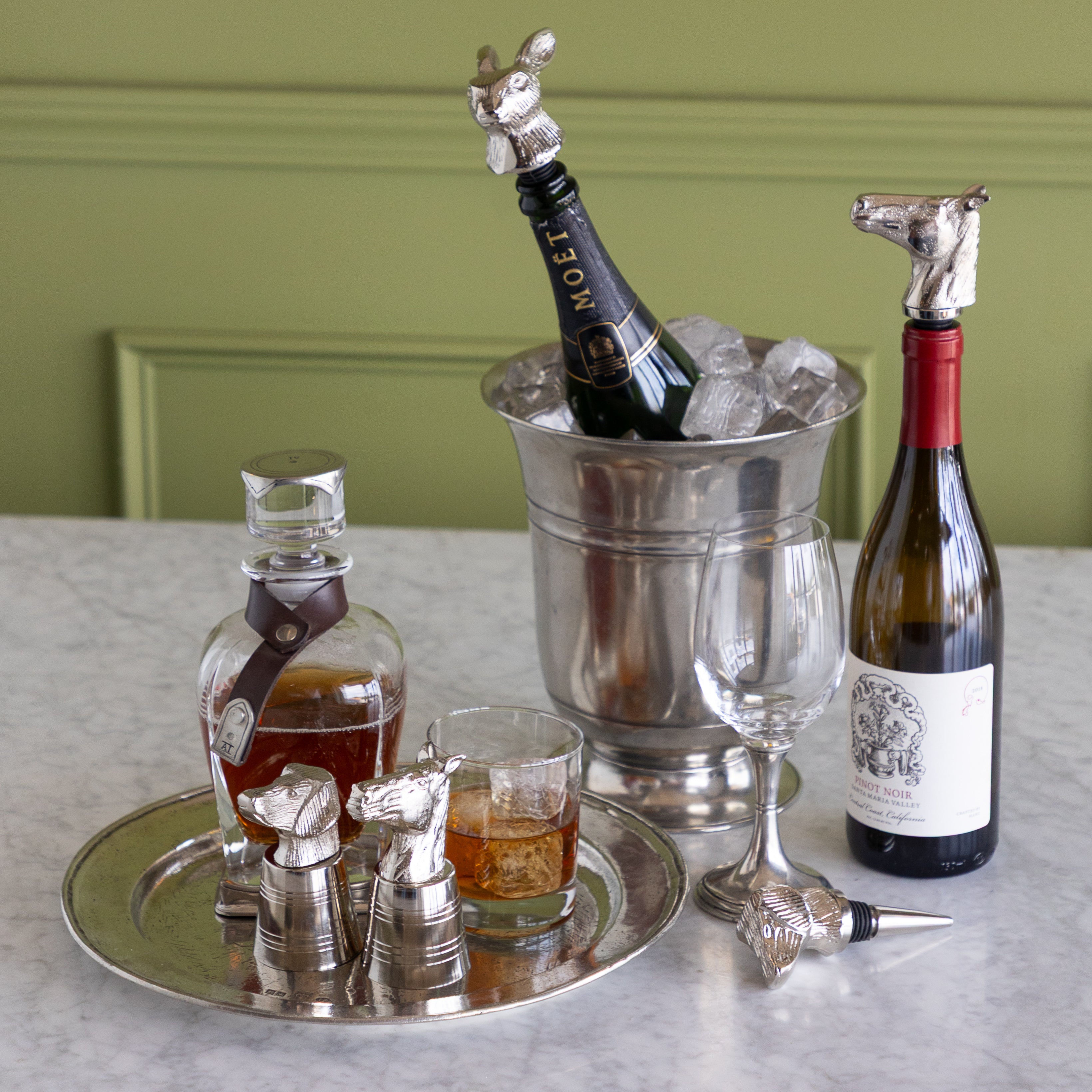 A styled bar or cocktail accessory set arranged on a marble surface against a green paneled wall. The set includes silver horse-shaped bottle stoppers, a horse-shaped decanter stopper, a horse-shaped ice bucket topper, a horse-shaped glass stopper, a horse-shaped cocktail relifer, and a horse-shaped bottle opener on a silver tray, alongside a wine bottle, a champagne bottle in an ice bucket, a wine glass, and a whiskey glass with amber liquid.