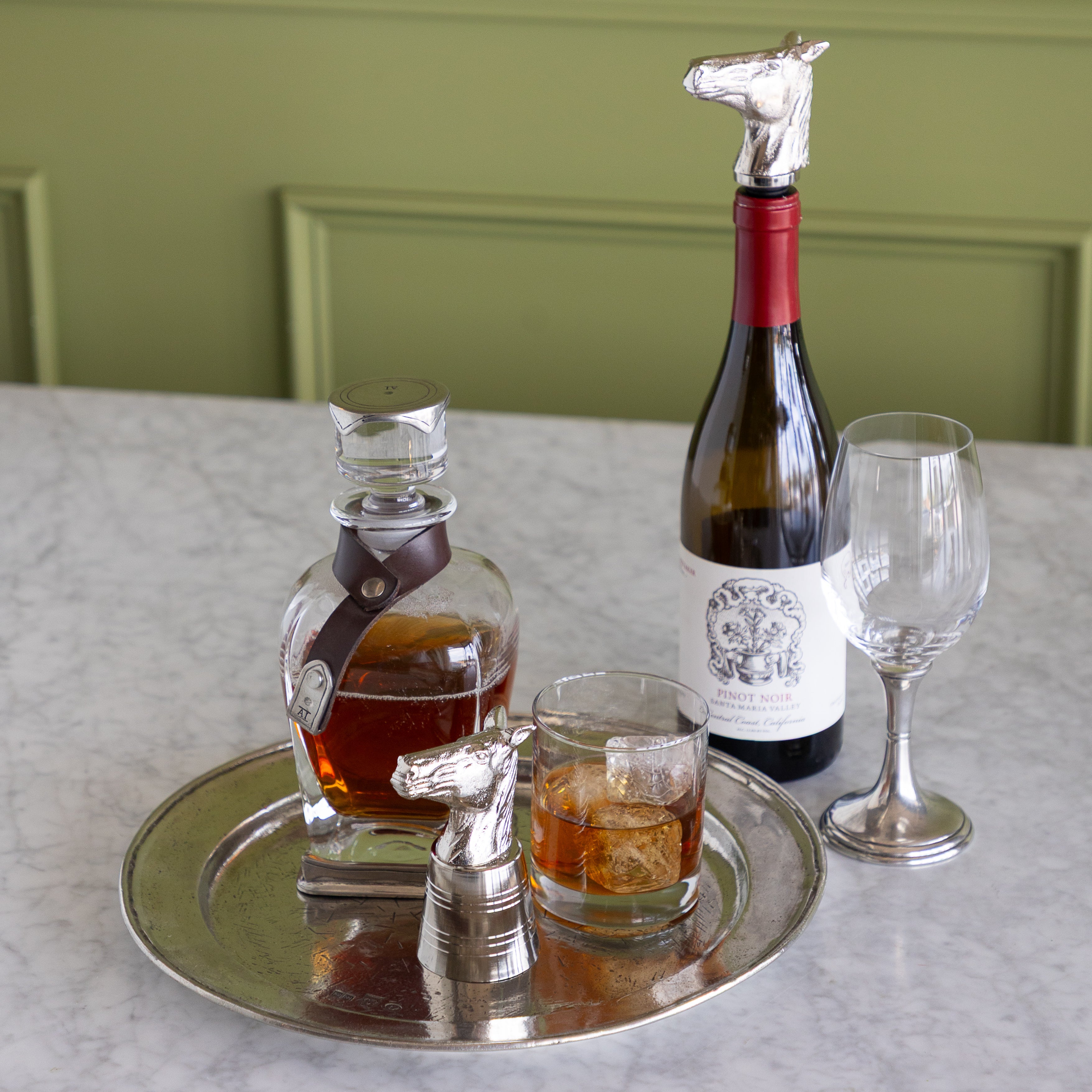 A styled tabletop scene with a silver tray holding a whiskey decanter, a filled rocks glass, an empty wine glass, and a wine bottle topped with a horse-shaped silver stopper. The background shows a pale green paneled wall and a light marble surface.