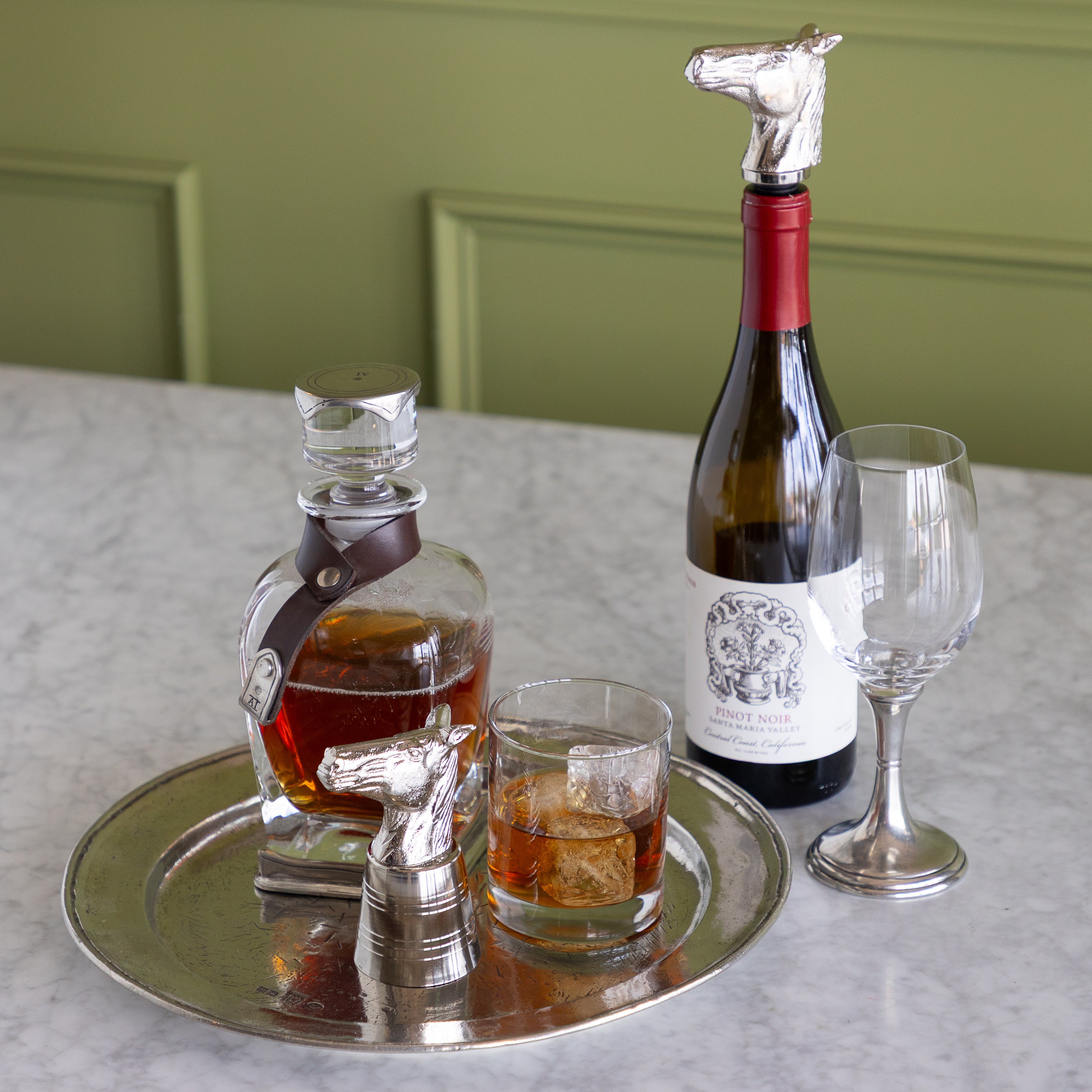 A styled tabletop scene with a silver tray holding a whiskey decanter, a whiskey glass, a wine glass, and a wine bottle topped with a horse-shaped silver stopper. The setting is indoors against a green paneled wall and light stone surface.