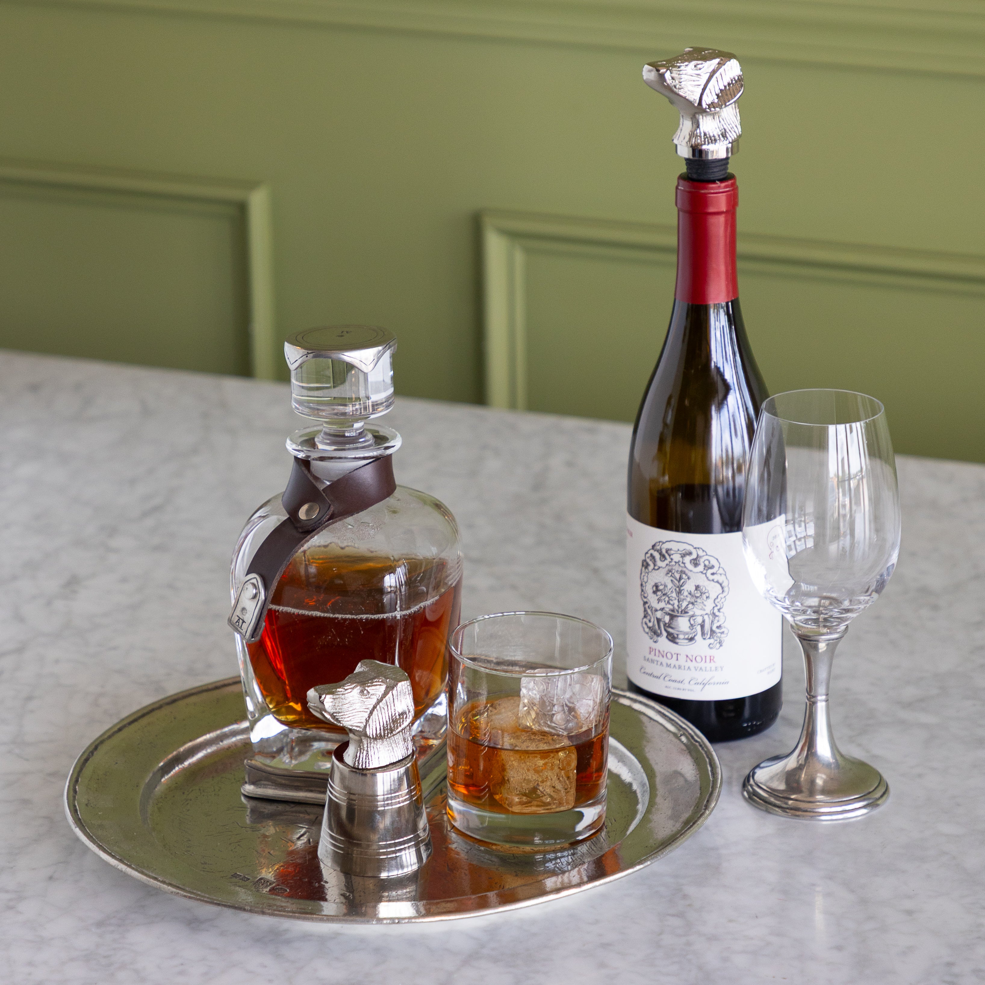 A silver-toned metal bottle stopper set arranged on a round silver tray on a marble table, alongside a wine bottle with a red foil top, an empty wine glass, and a small glass of amber liquid. The background shows olive green paneled walls.