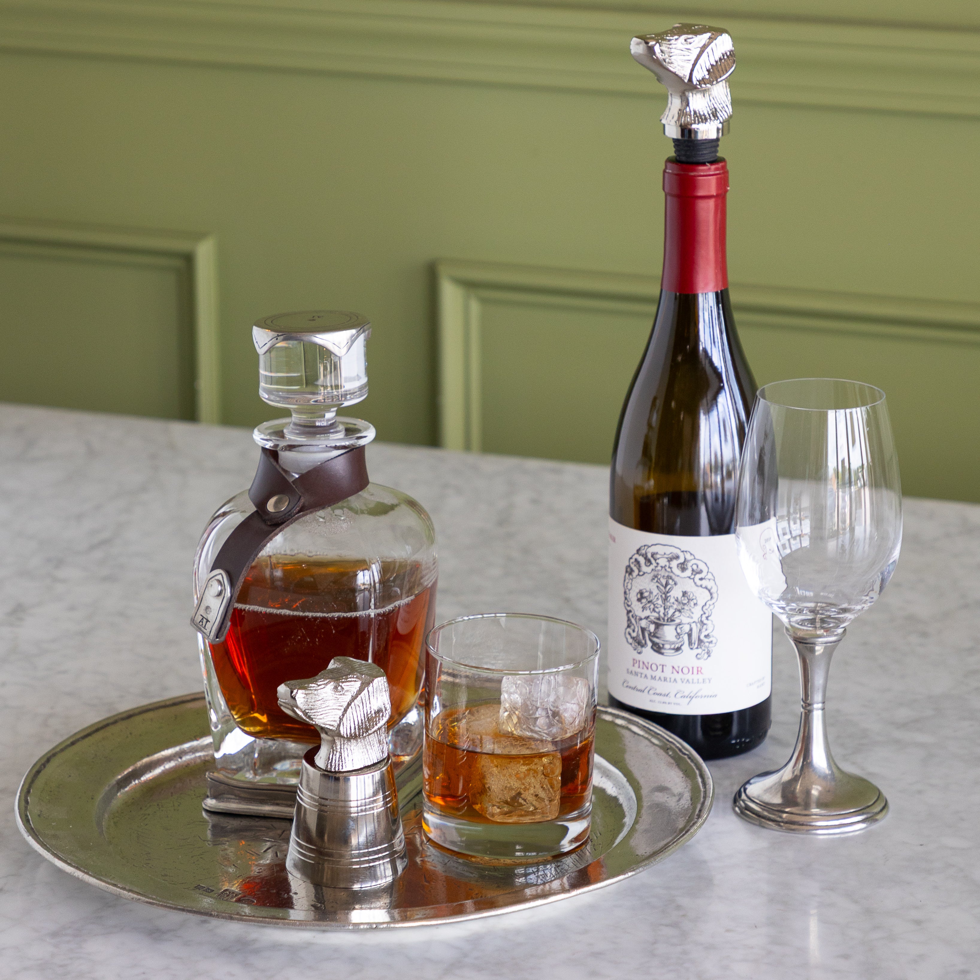 A styled still life on a marble countertop featuring a silver tray with a clear glass decanter of amber liquor, a small silver jigger, a filled rocks glass, and a wine bottle with a foil stopper next to a stemmed wine glass. The background shows a sage green paneled wall.
