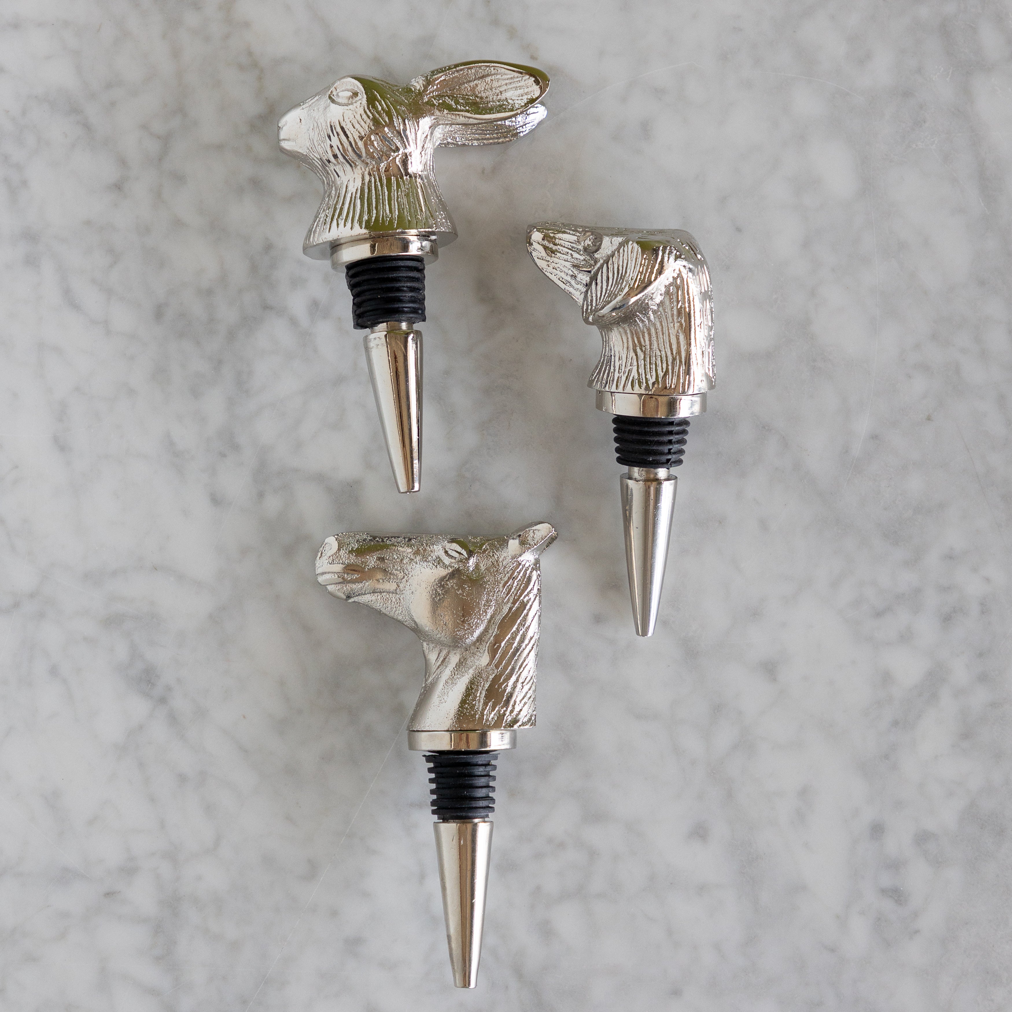 Three silver-toned metal wine bottle stoppers shaped like animal heads, including a rabbit, a duck, and a horse, arranged on a light gray marble surface.