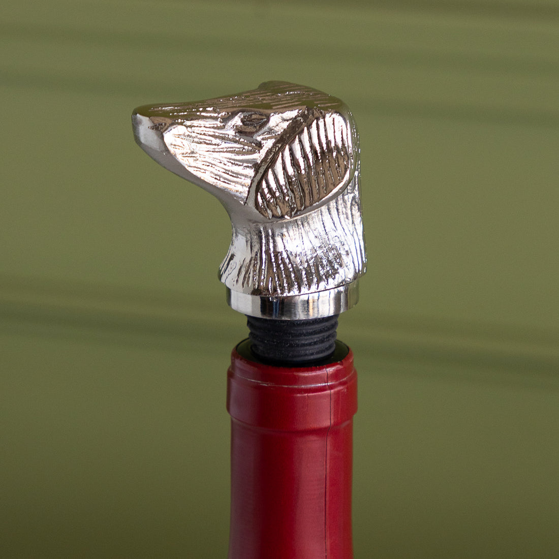 A close-up of a wine bottle featuring a polished silver metal dog bottle stopper, set against a soft olive green background.