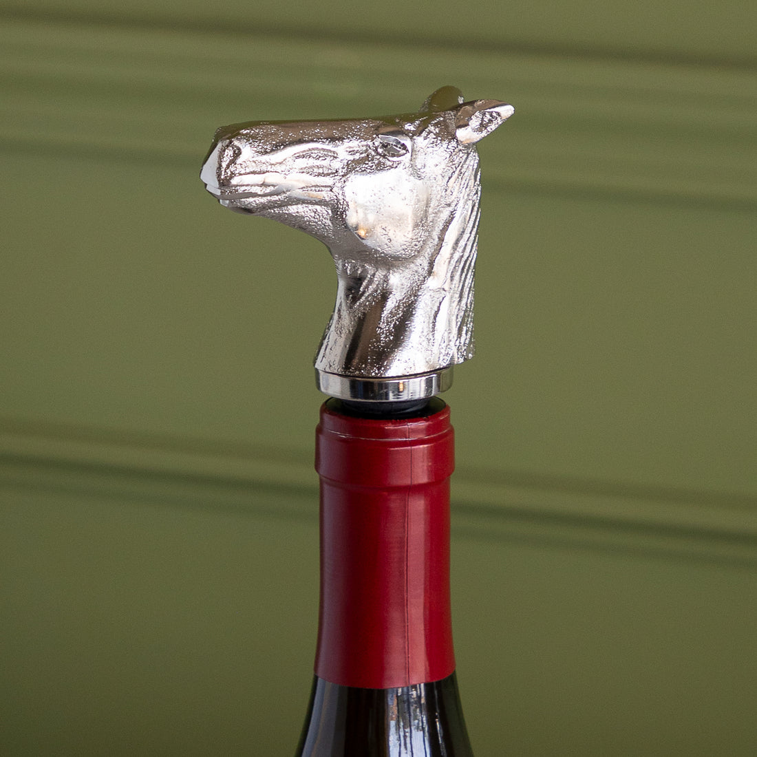 A silver metallic horse-shaped bottle opener mounted on the neck of a wine bottle with a red foil capsule, set against a soft olive green background.