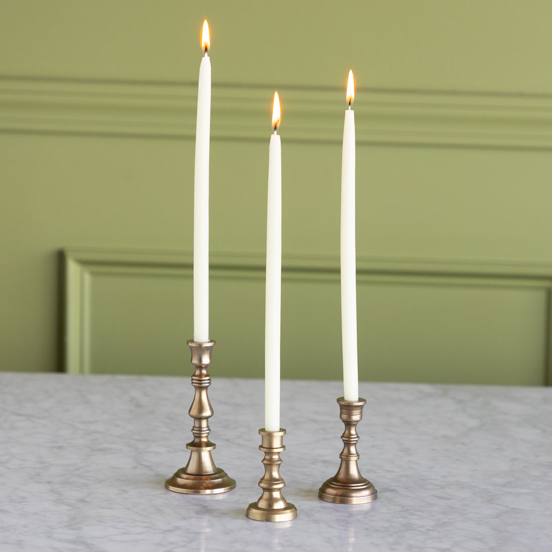 Three white candles in bronze candlesticks on a marble surface with a green paneled wall background.