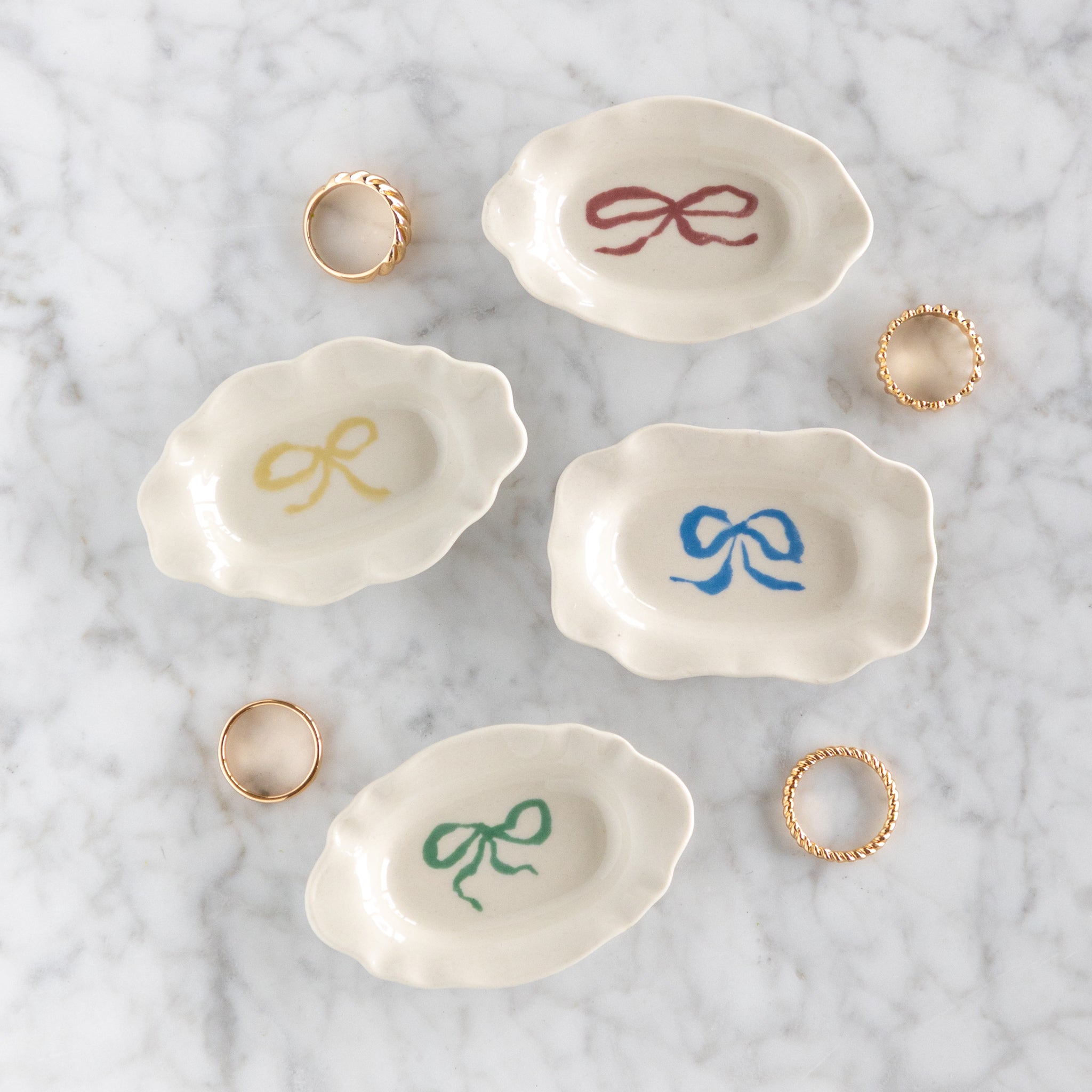 Set of four small ceramic dishes with colorful bow designs on a marble surface with rings.