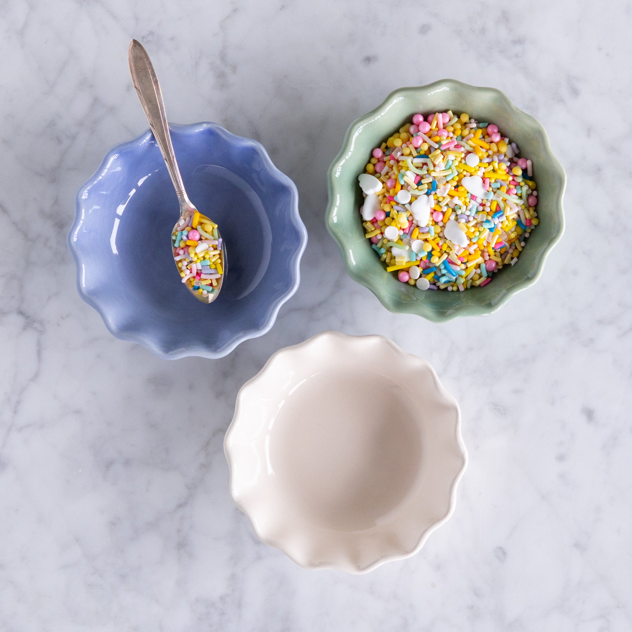 Three ceramic bowls in green, white and cream with colorful sprinkles on a marble surface.