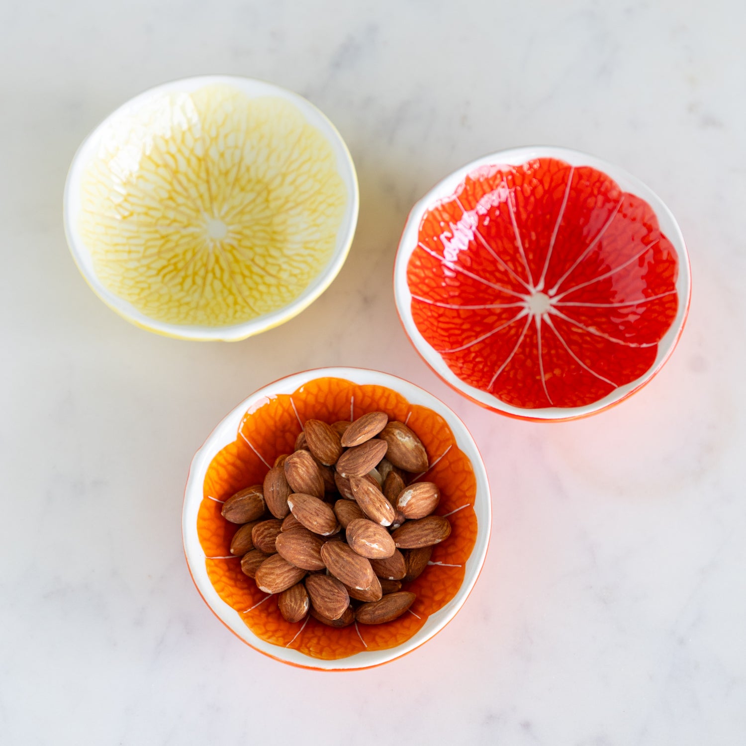 Three Ceramic Citrus Fruit Shaped Bowls ––one filled with almonds on a marble table.