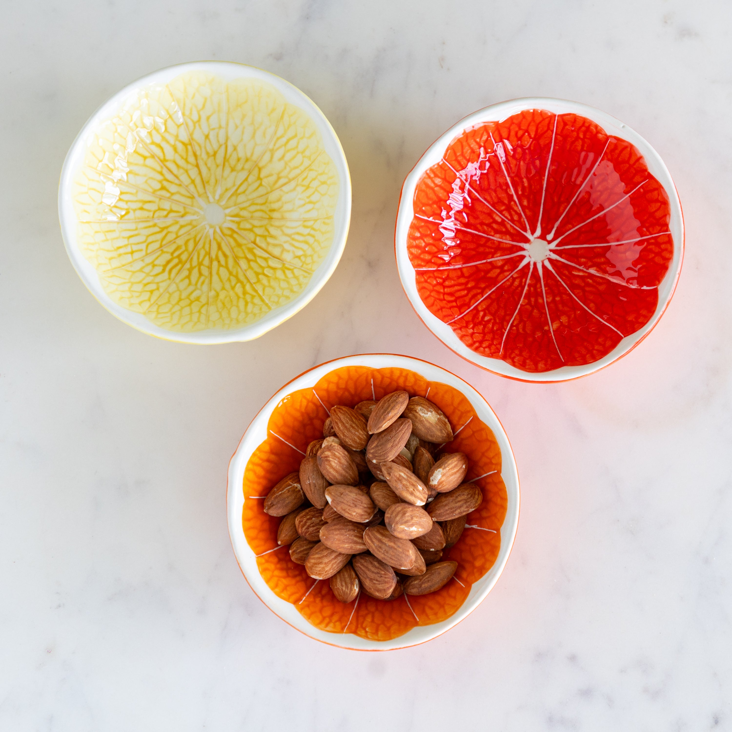 Three Ceramic Citrus Fruit Shaped Bowls, one with almonds, on a marble table.