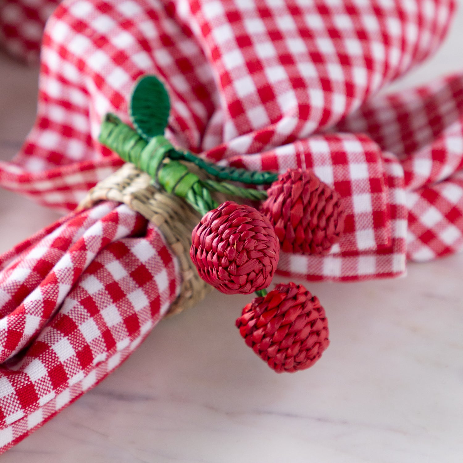 Decorative napkin ring with red cherries and green stem on a red and white checkered cloth.
