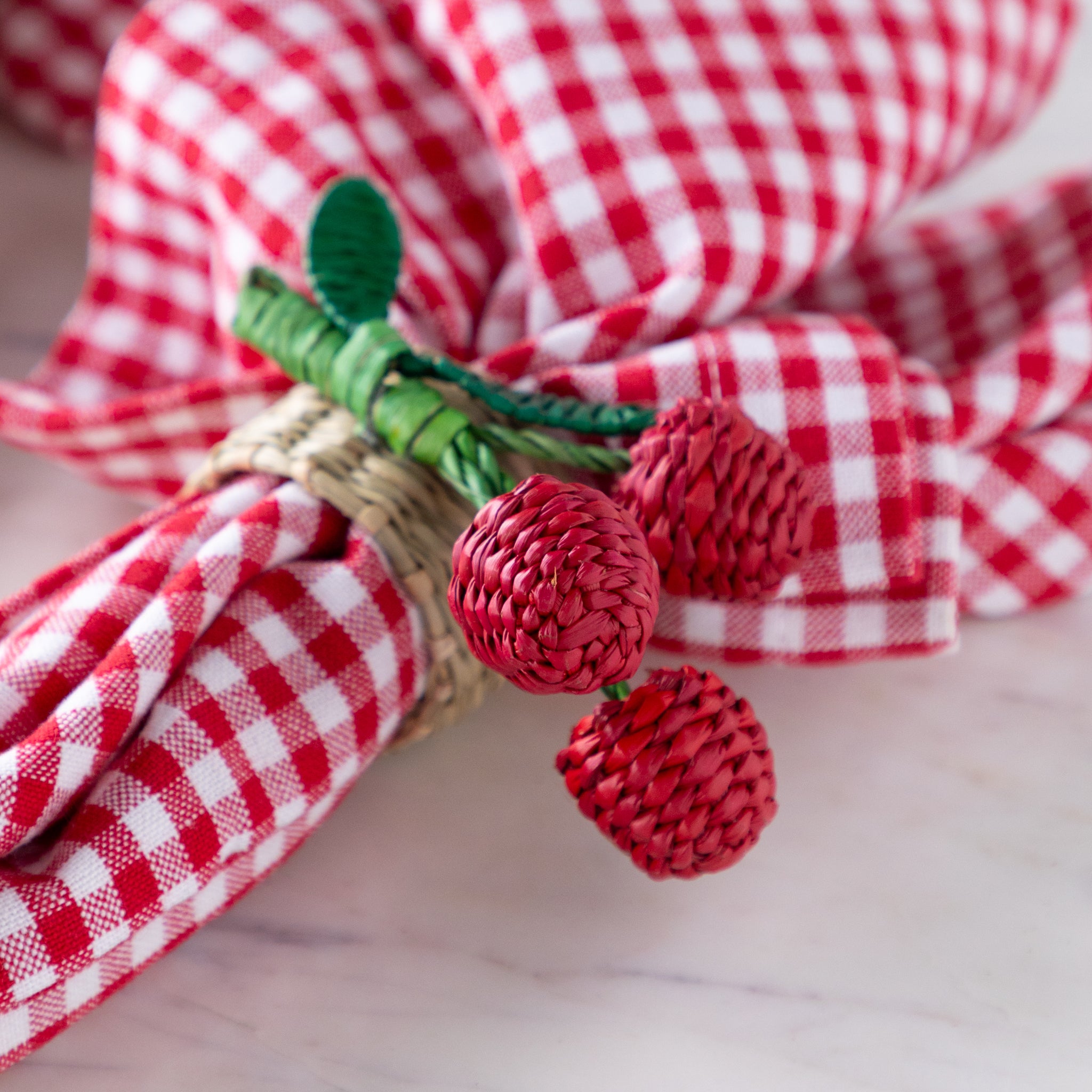 Decorative napkin ring with red cherries and green stem on a red and white checkered cloth.
