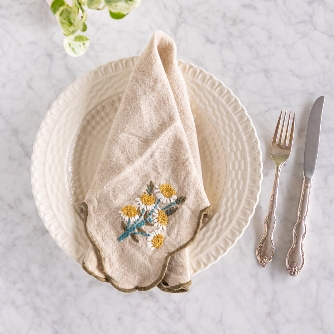 Beige embroidered daisies napkin on a white plate with silverware on a marble surface.