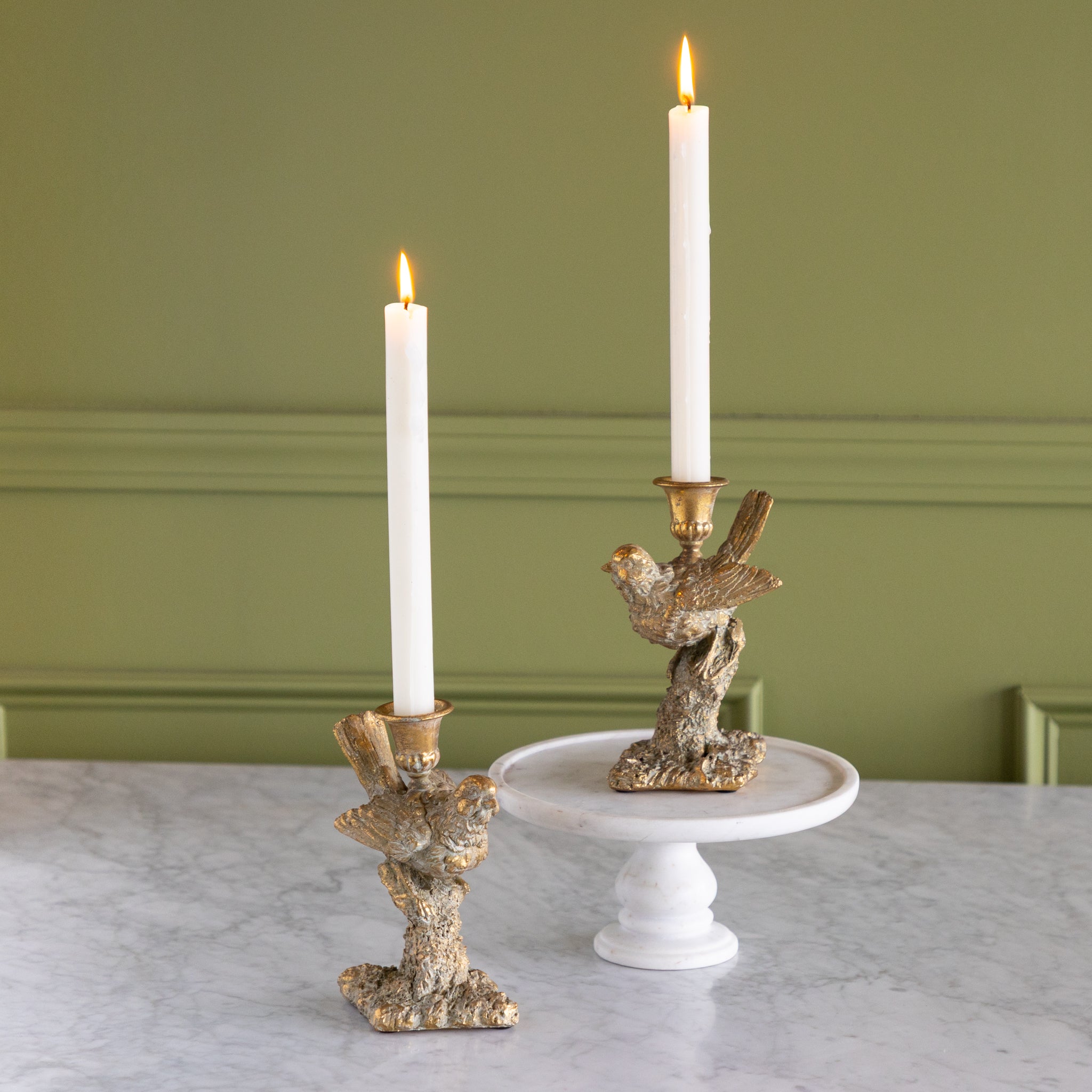 Two Bird Shaped Taper Holders with lit candles on a marble table and green wall background.