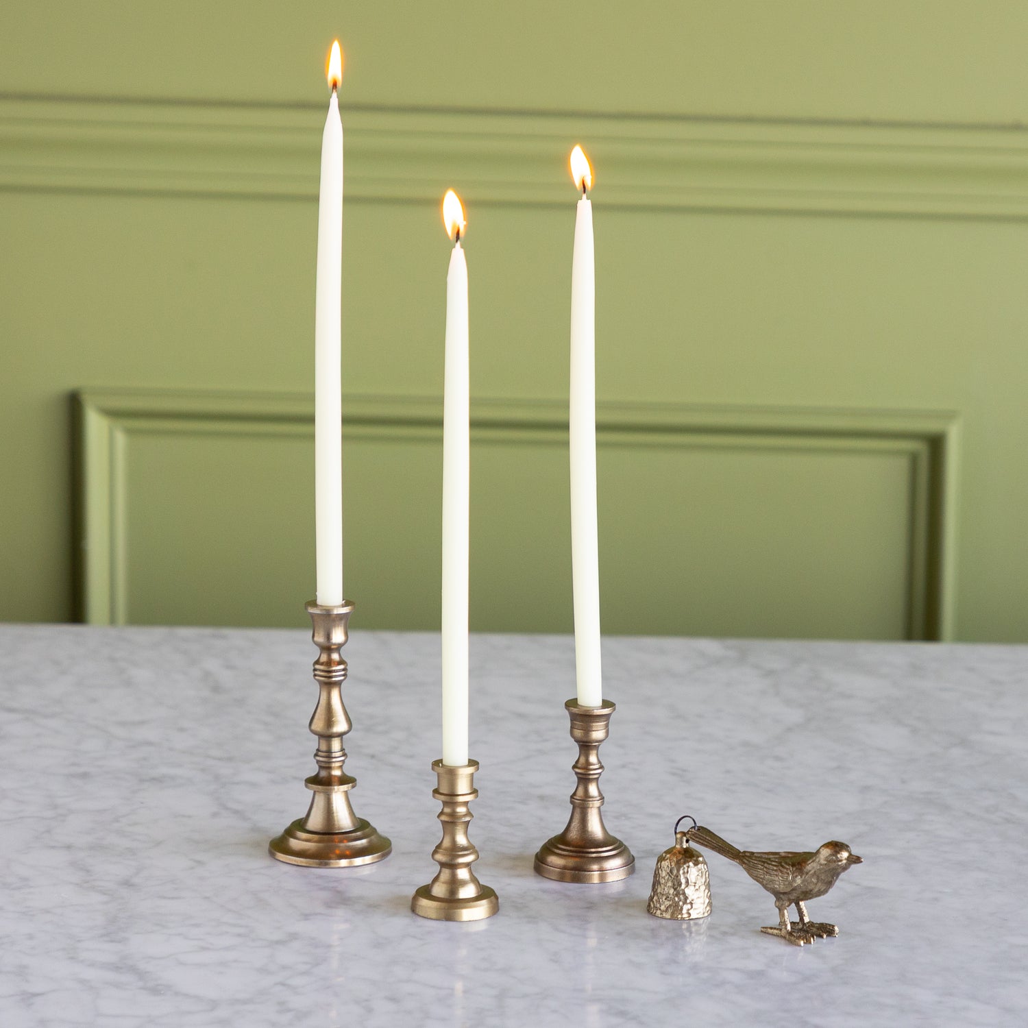 Three lit candles in brass candlesticks on a marble surface with a green wall background.