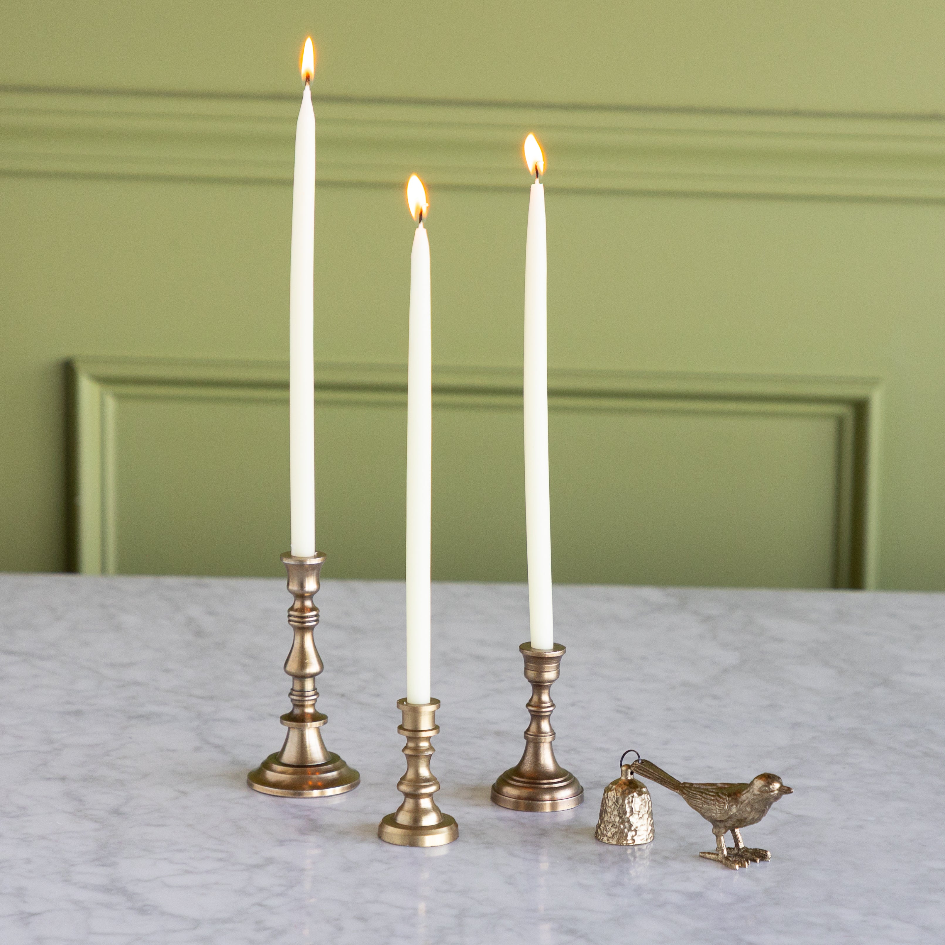 Three lit candles in brass candlesticks on a marble surface with a green wall background.