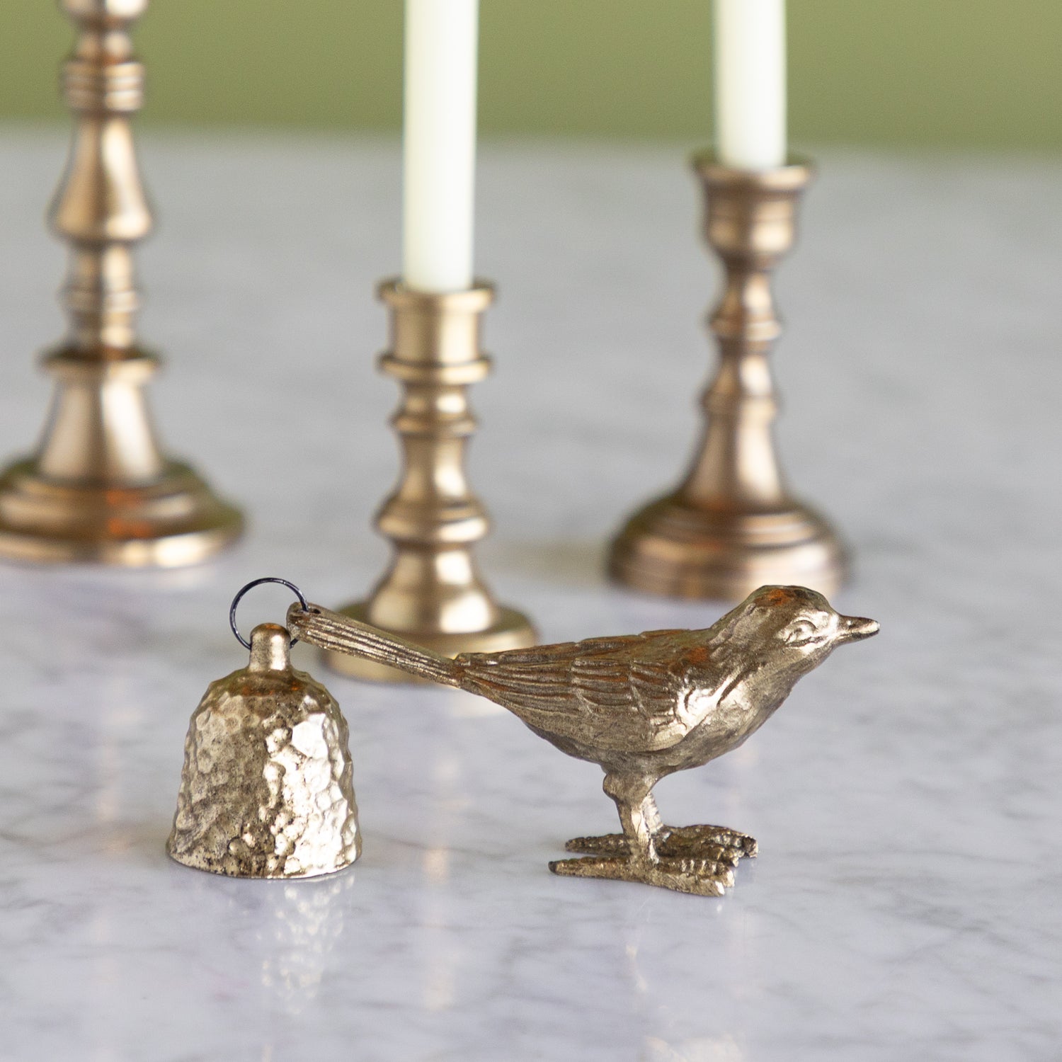 Bronze bird-shaped bell candle snuffer on a marble surface with a blurred green background.