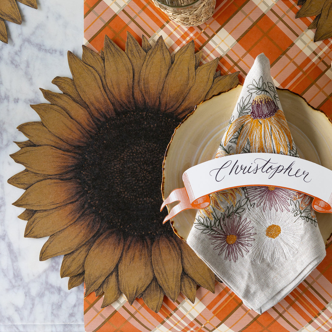 A fall place setting with the Die-cut Vintage Sunflower Placemat, wood slice plate with the Autumn Mums Napkin and Classic Orange Banner Table Accent atop, on the Orange Plaid Runner.