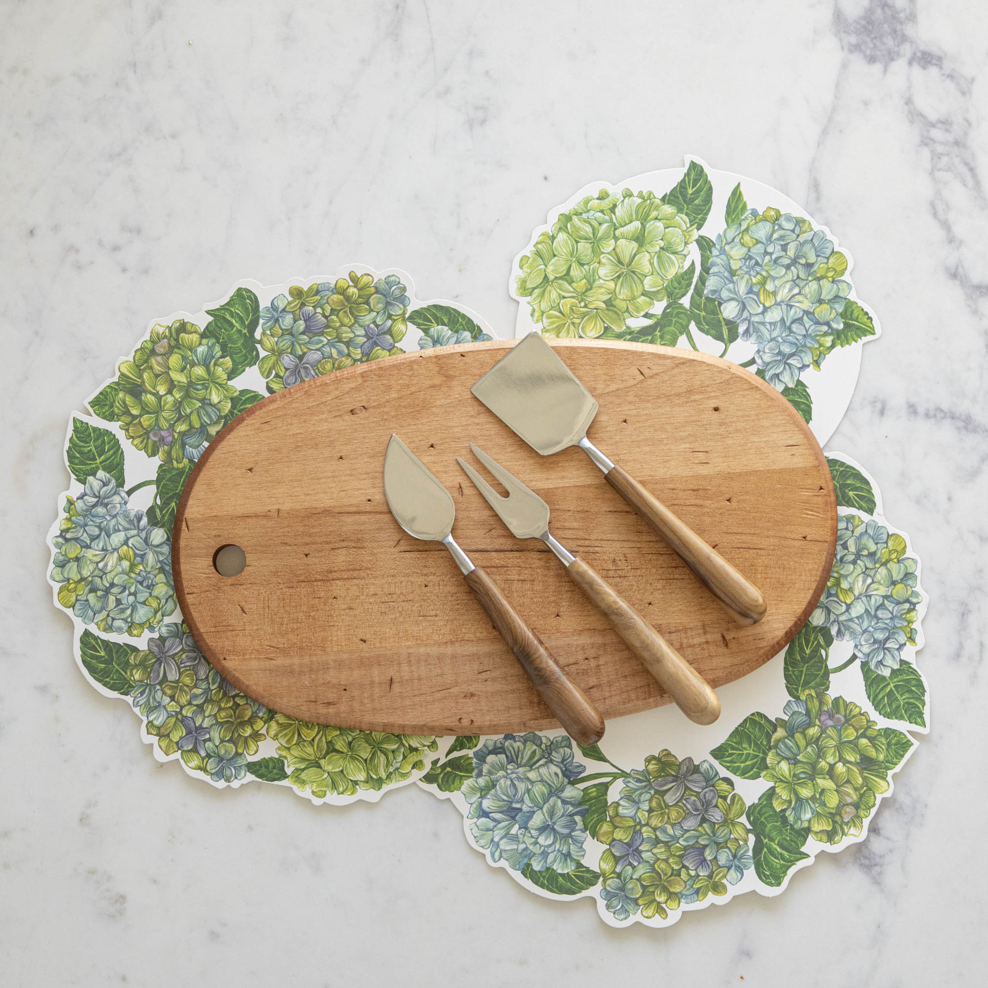 The Maple Oval Serving Board with a Teak &amp; Stainless Cheese Server Set on top, with Hydrangea Serving Papers underneath.