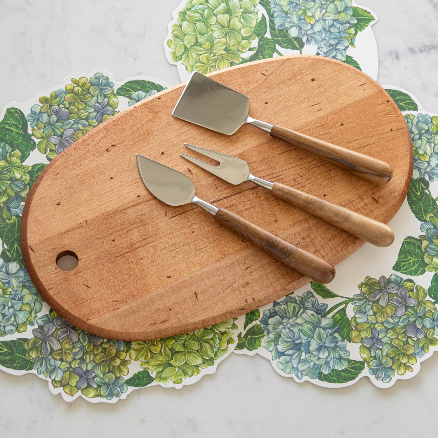 The Maple Oval Serving Board with a Teak &amp; Stainless Cheese Server Set on top, with Hydrangea Serving Papers underneath.