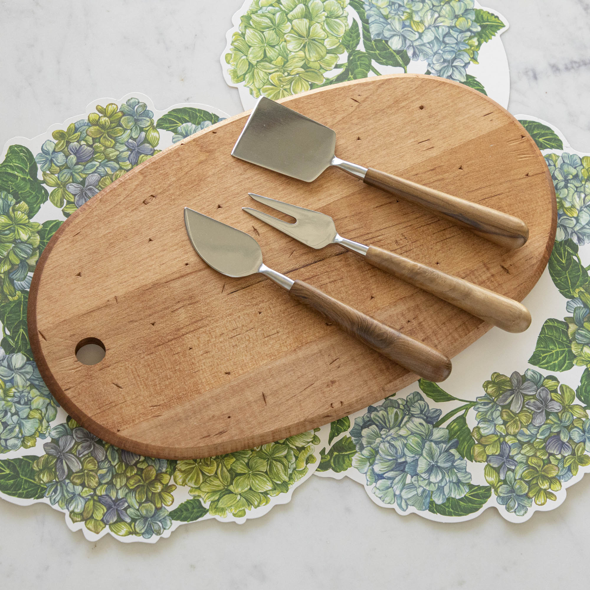 The Maple Oval Serving Board with a Teak &amp; Stainless Cheese Server Set on top, with Hydrangea Serving Papers underneath.