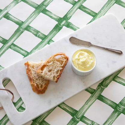 A White Marble Cheese Board with sliced bread and butter and a butter spreader on top, on top of a green lattice runner.