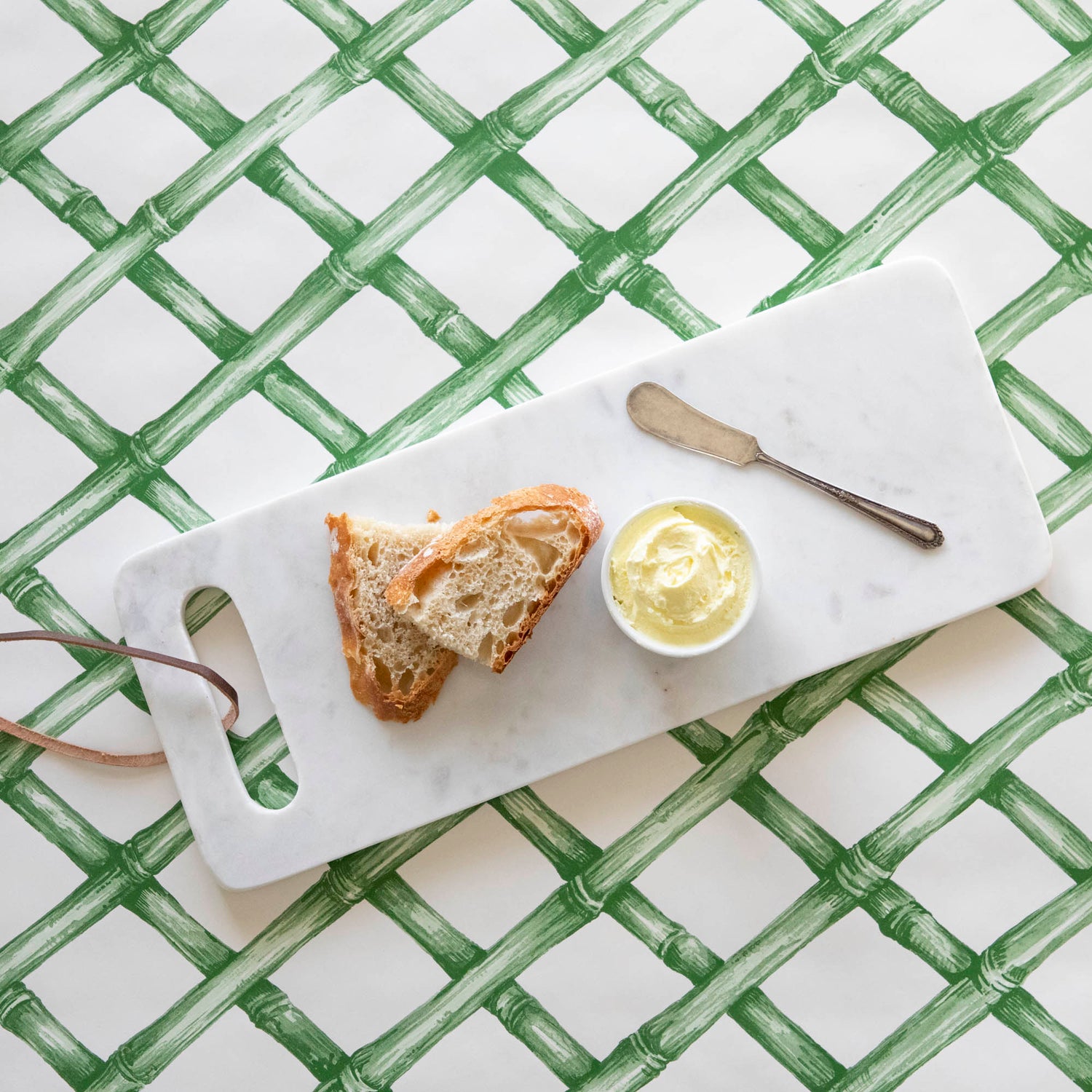 A White Marble Cheese Board with sliced bread and butter and a butter spreader on top, on top of a green lattice runner.