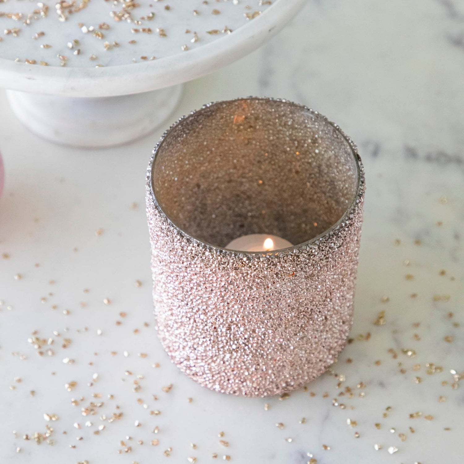 Pink Shimmer Votive surrounded by glitter with a lit tea light candle in the middle.