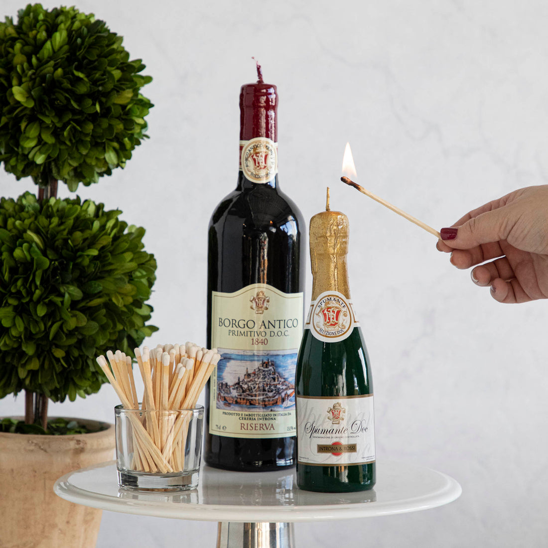 Lighting a high-quality Cereria Introna Spumante Piccola Shaped Candle on a wine bottle with a match, alongside another bottle and matches on a white stand.