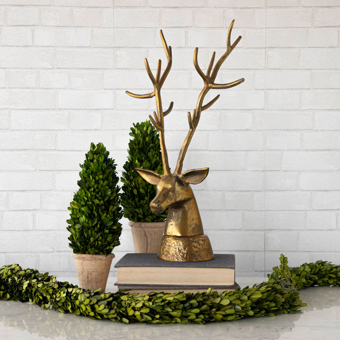 A rustic, metal Deer head on a stack of books, surrounded by boxwood.