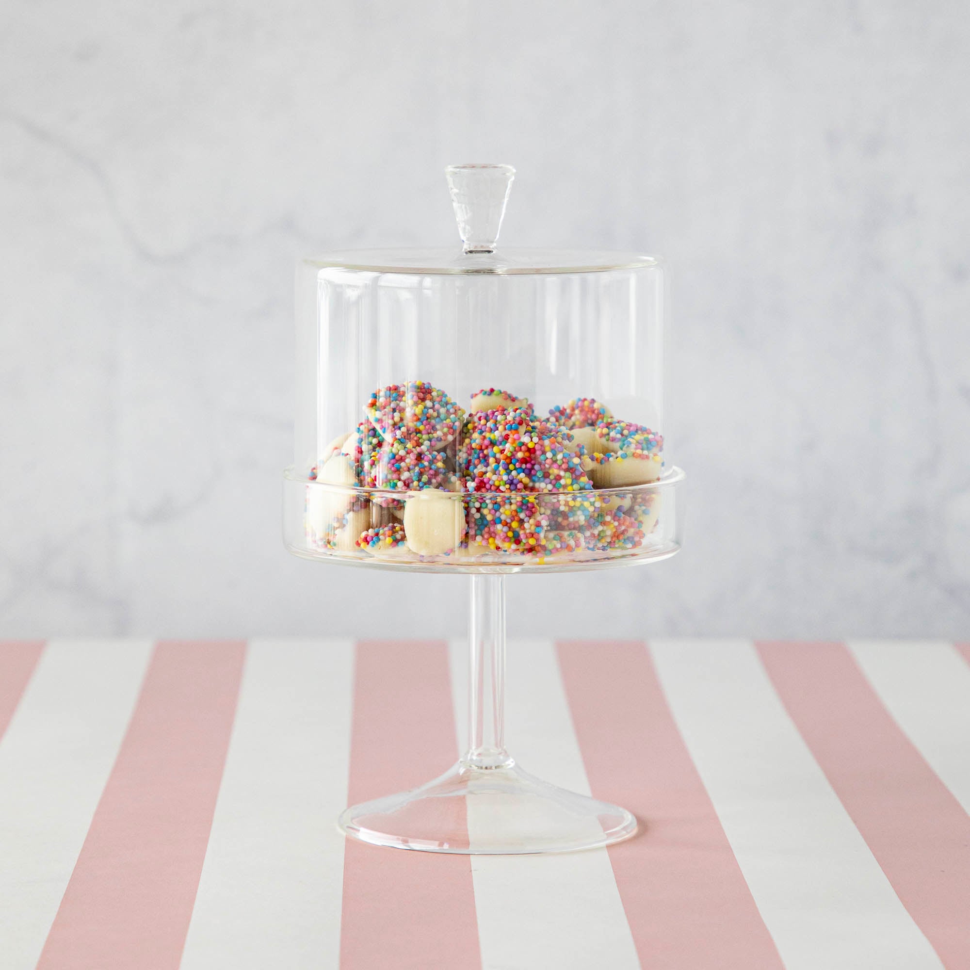 A HomArt Glass Dome &amp; Pedestal cake stand with a glass dome lid filled with sprinkle-covered cookies on a pink and white striped surface.