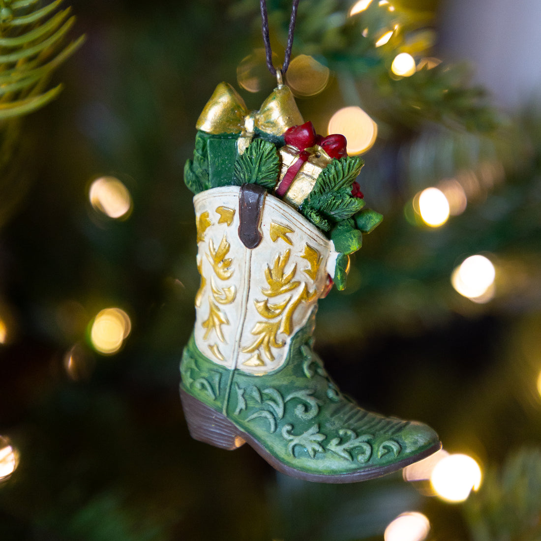 A Christmas ornament shaped like a green cowboy boot with gold western embroidery, hanging in front of a blurred Christmas tree with warm white lights.