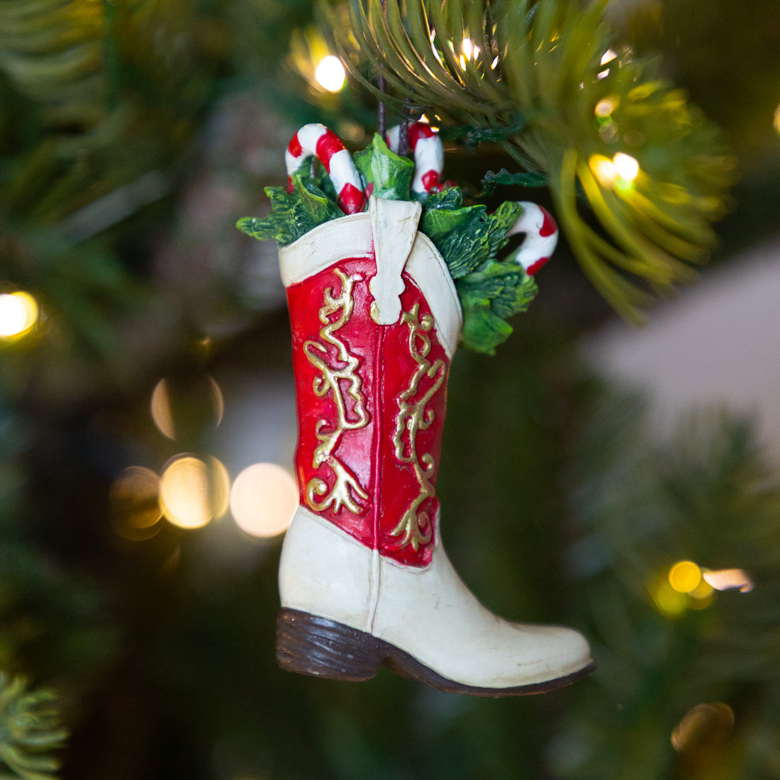 A Christmas ornament shaped like a cowboy boot hanging on a Christmas tree. The boot is cream-colored with a bright red shaft decorated with gold western-style embroidery, and green pine branches with candy canes peeking out from the top.