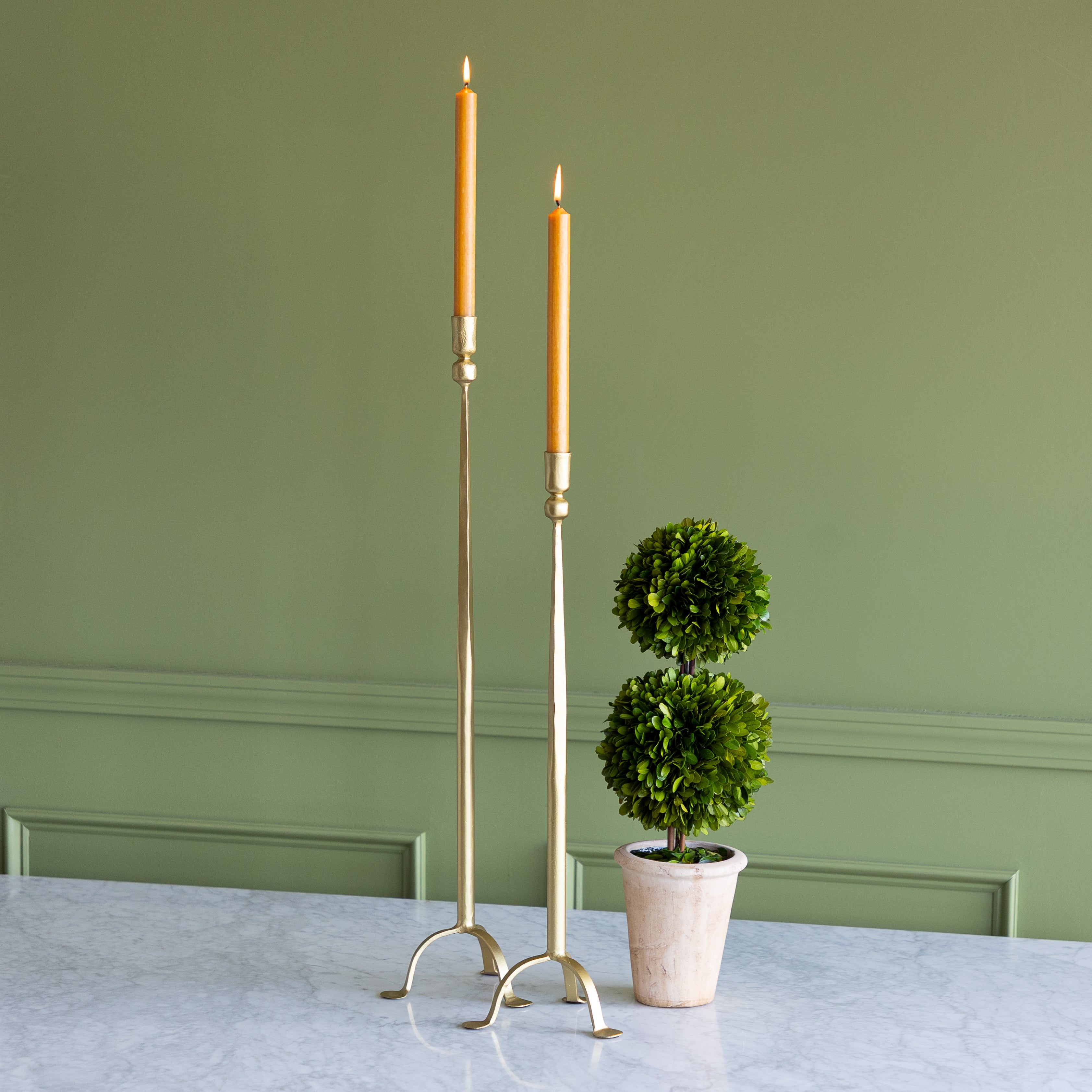 Two elongated golden candlesticks with lit orange candles on a green background. 