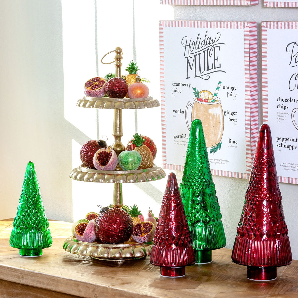 Decorative Christmas trees and a tiered fruit stand on a wooden surface with a 'Holiday Mule' recipe in the background.
